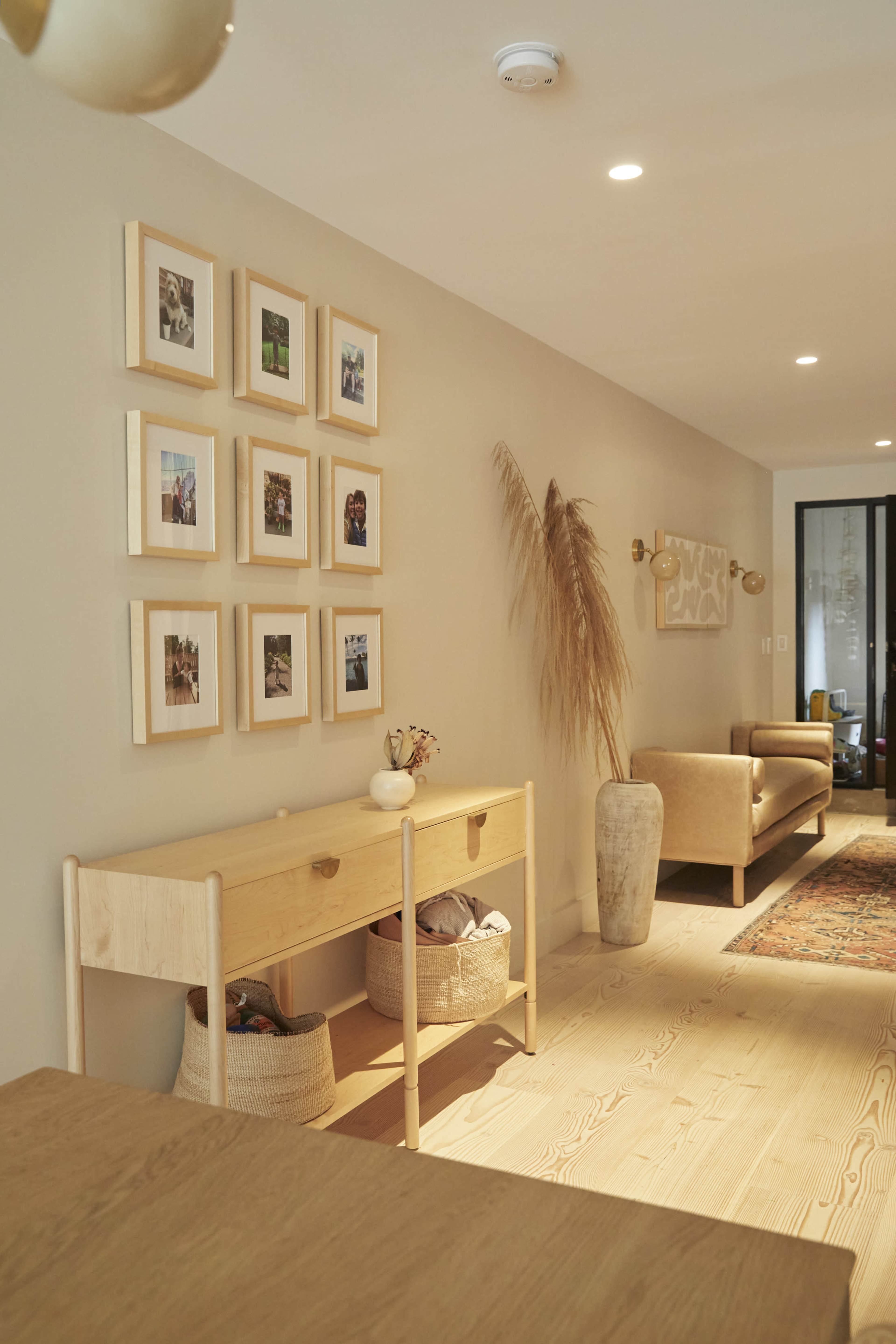 A well-lit hallway featuring a wooden console table, framed photographs on the wall, and a beige sofa, with a decorative vase and dried plants.