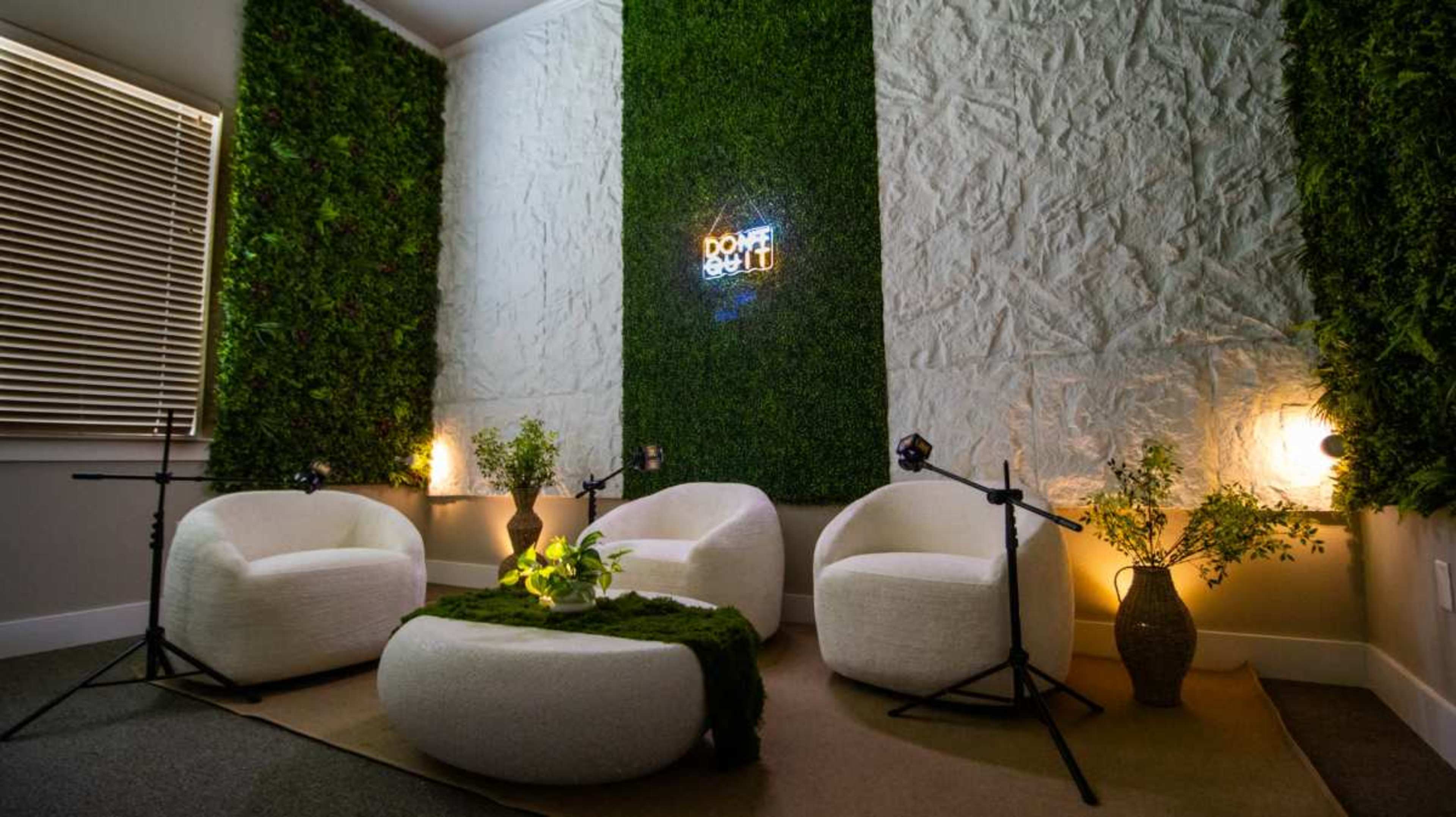 The image shows a cozy room with green, moss-covered walls, three white circular chairs surrounding a round table, and two plants in decorative vases, illuminated by soft lighting.