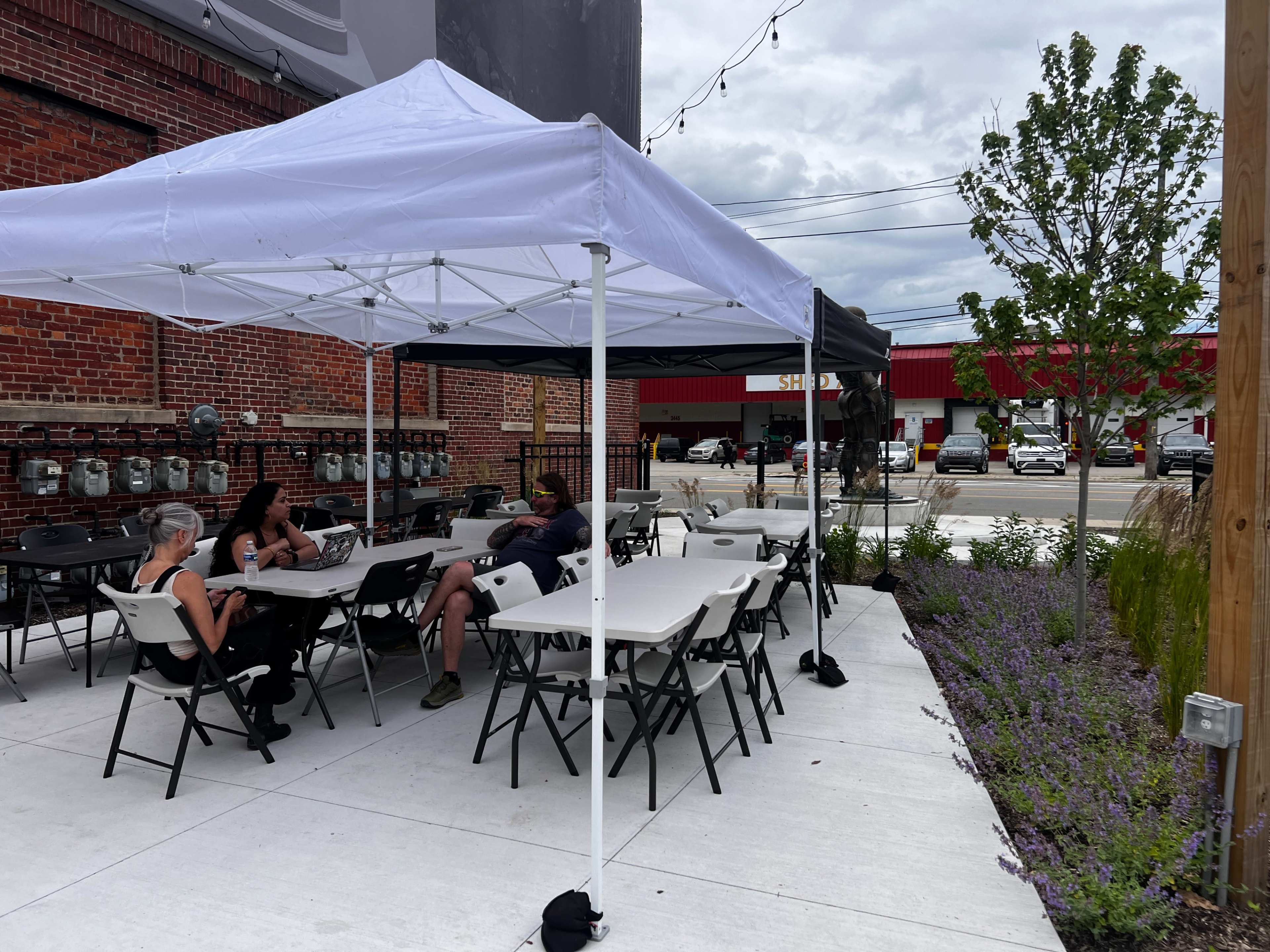 A shaded outdoor seating area is set up with tables and chairs, where people are sitting and working, surrounded by plants and a parking lot in the background.