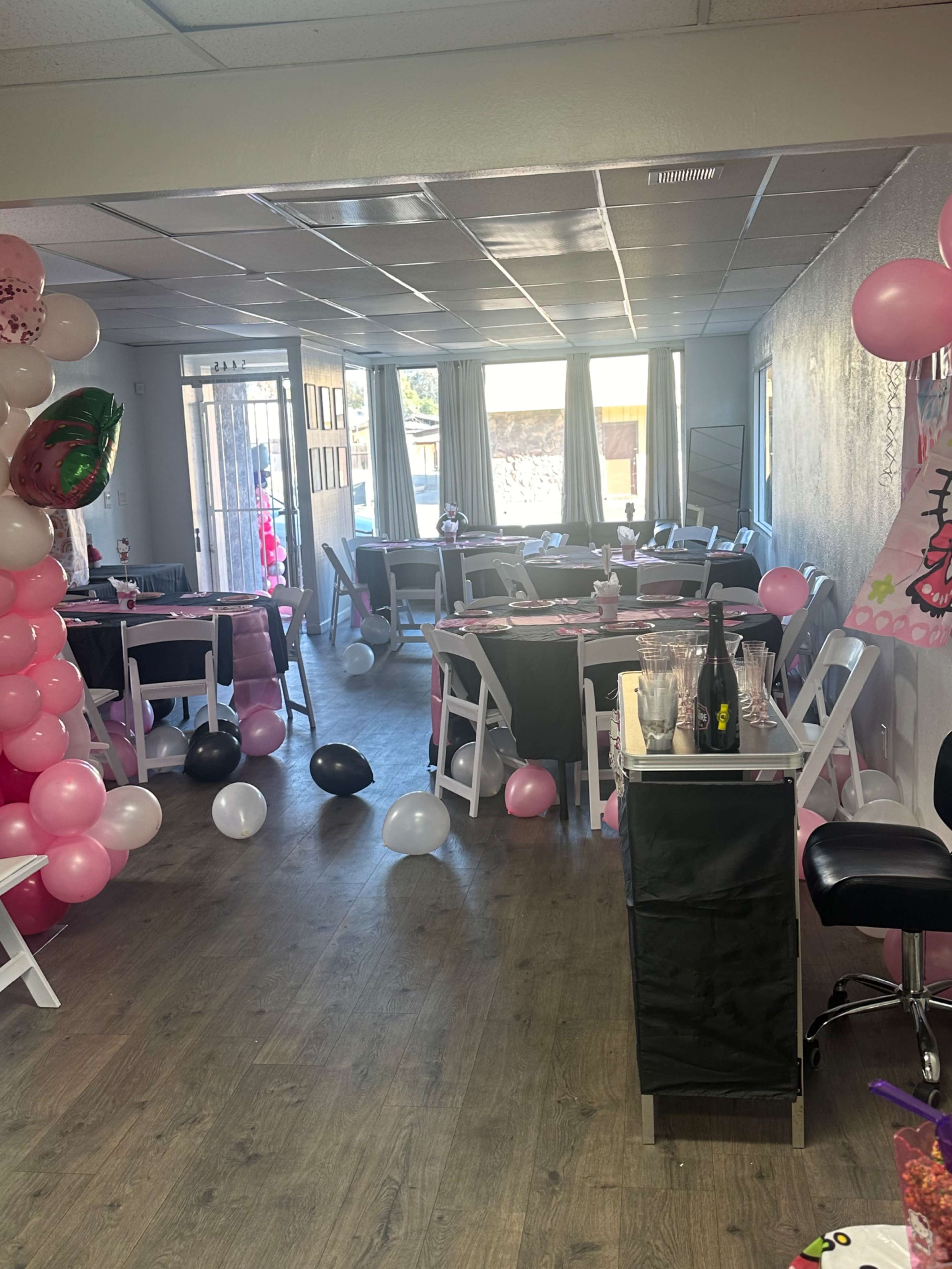The interior of a party venue features pink and black balloons, table settings with pink tablecloths, and a festive atmosphere.