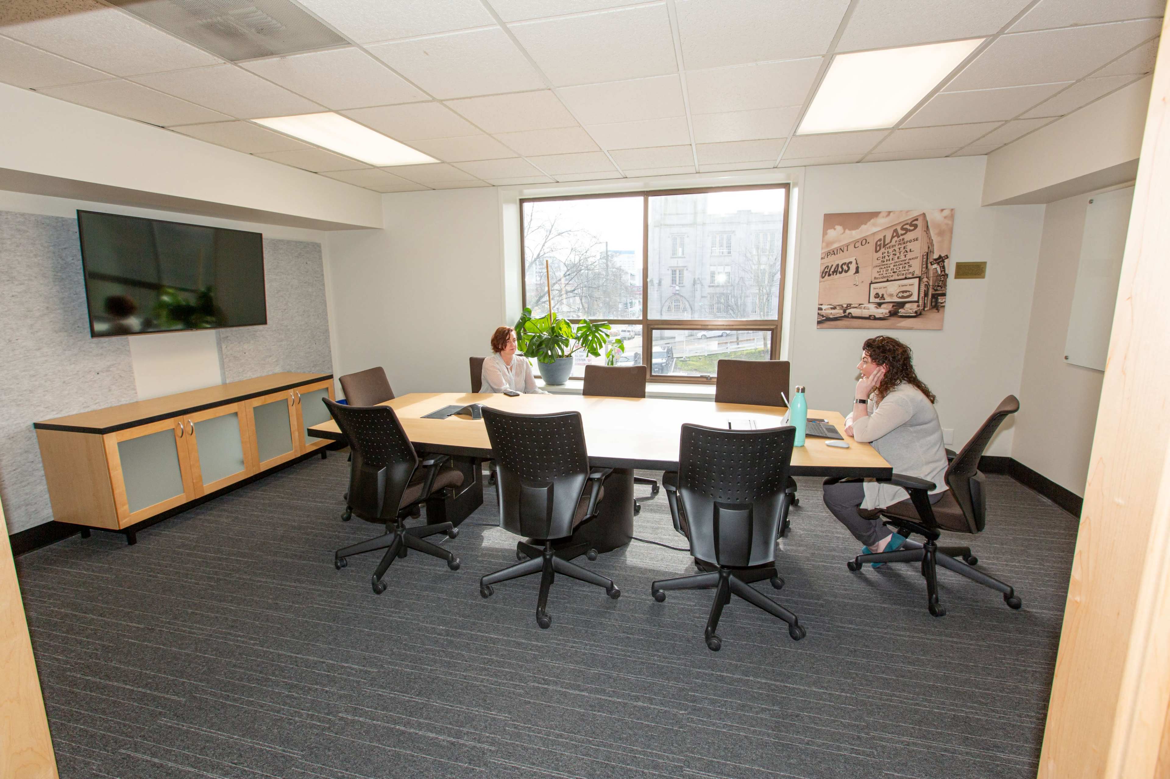 Two individuals are seated at a large rectangular table in a brightly lit conference room, with a television and a plant in the background.