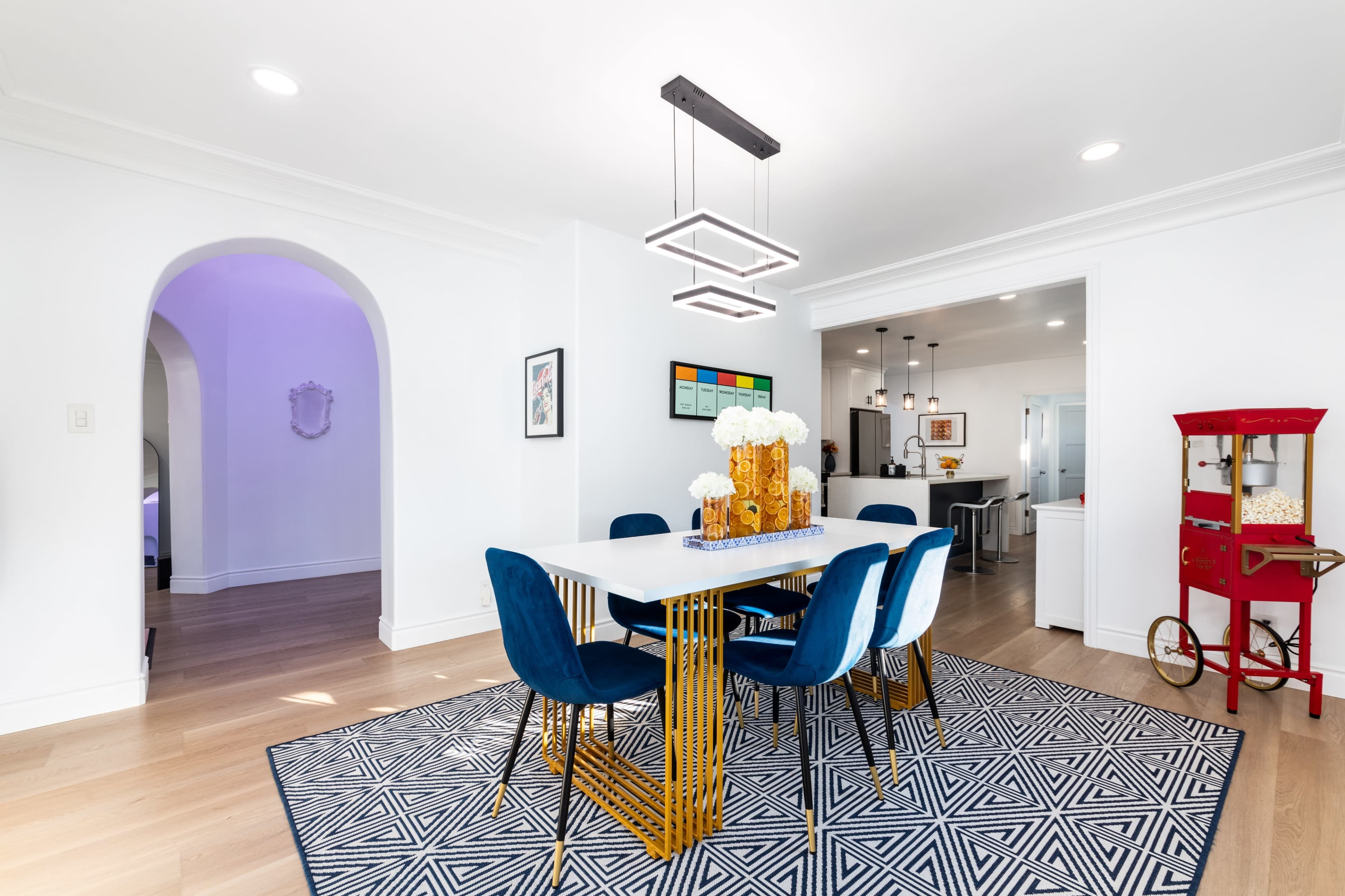A modern dining area features a white table surrounded by blue chairs, a geometric rug, and a decorative centerpiece, with an archway leading to another room.