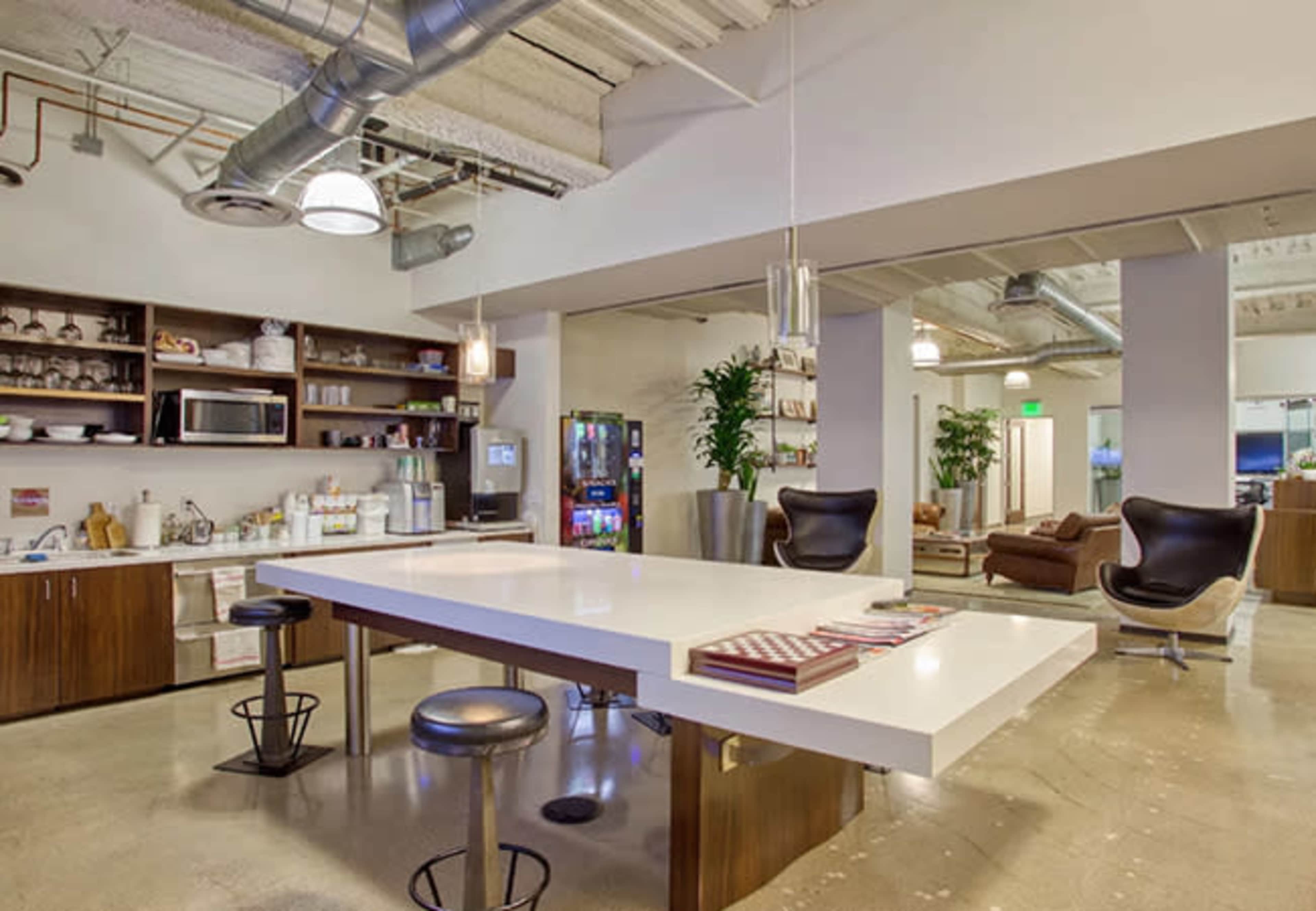 The image shows a modern office kitchen area featuring a long white table, bar stools, shelves with kitchenware, and a seating area with two chairs.