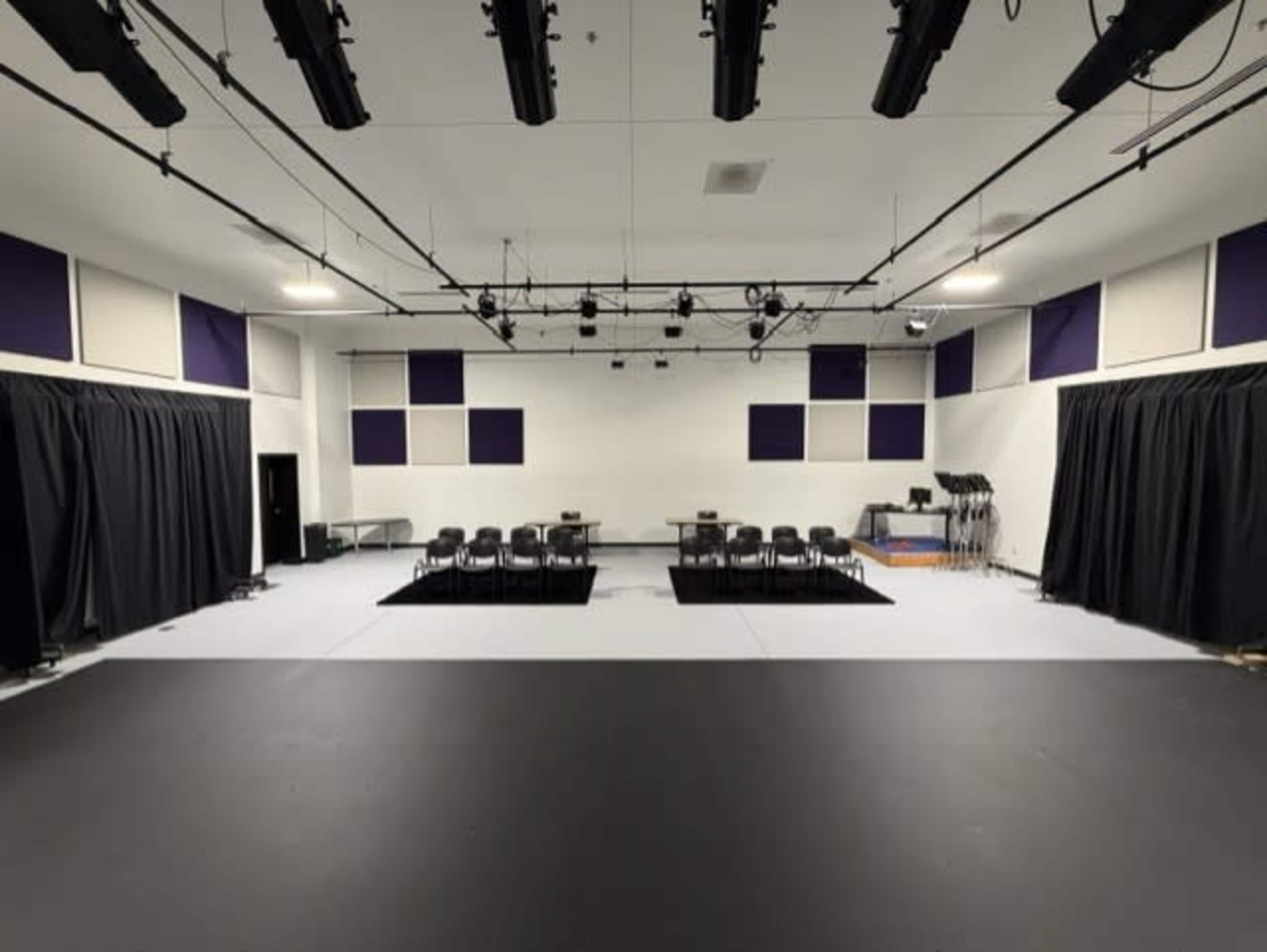 The image shows a spacious, well-lit rehearsal or performance studio with black curtains, a row of chairs, and a central table set up on a black mat.