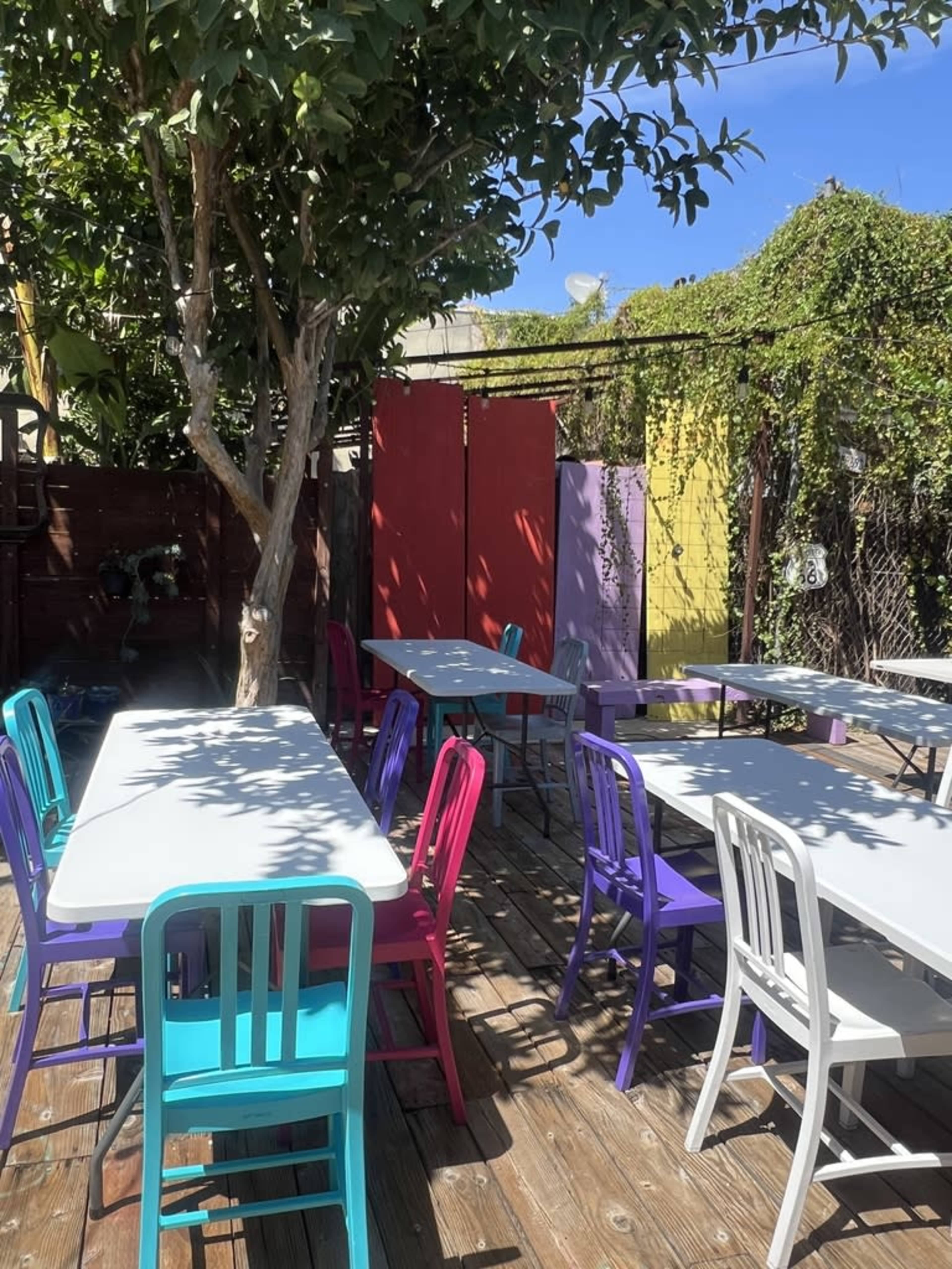 The image shows an outdoor seating area with several tables and colorful chairs surrounded by greenery and vibrant murals.