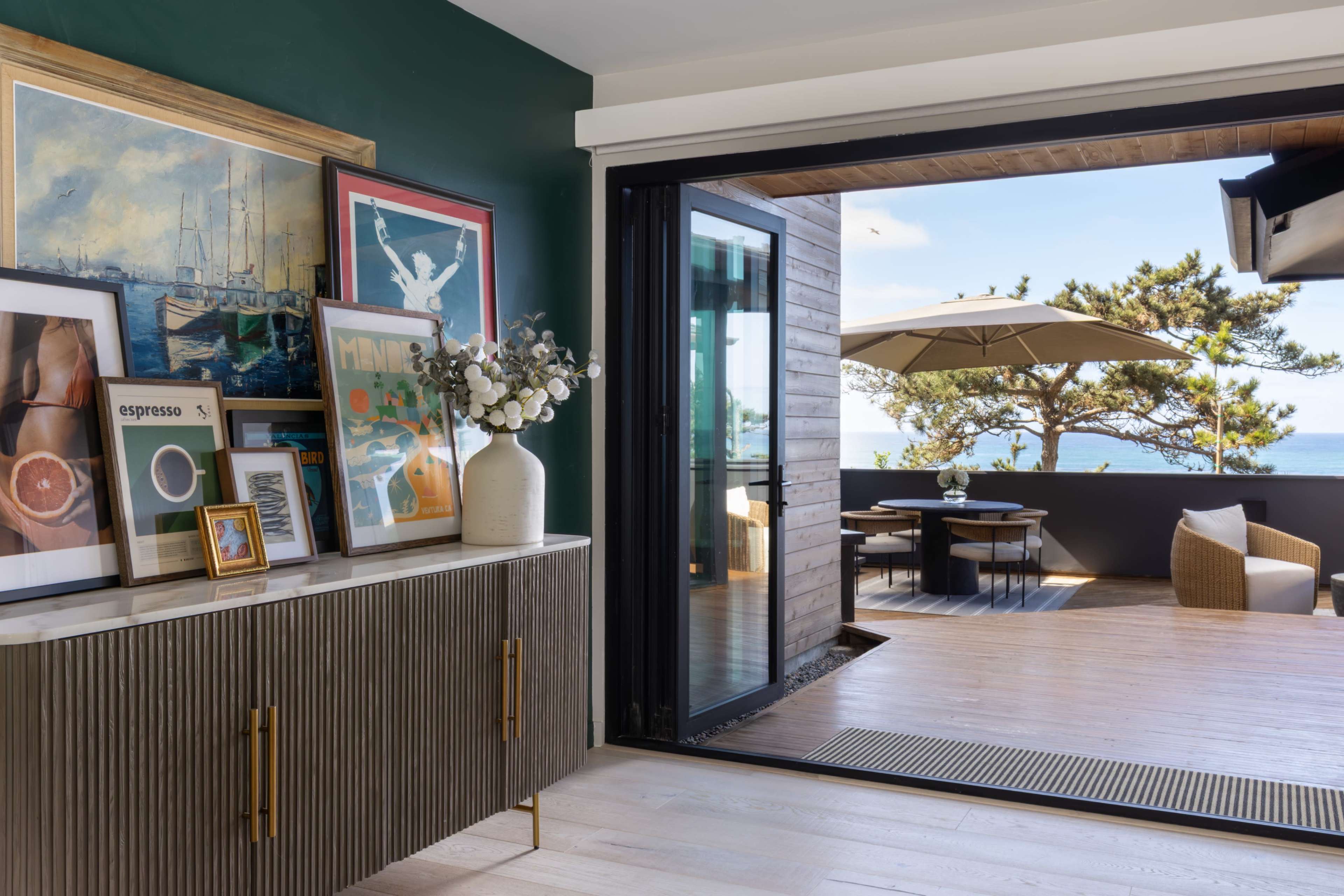 A modern living space features a console table adorned with framed artwork and a vase, opening up to a deck with outdoor seating and a view of the ocean.