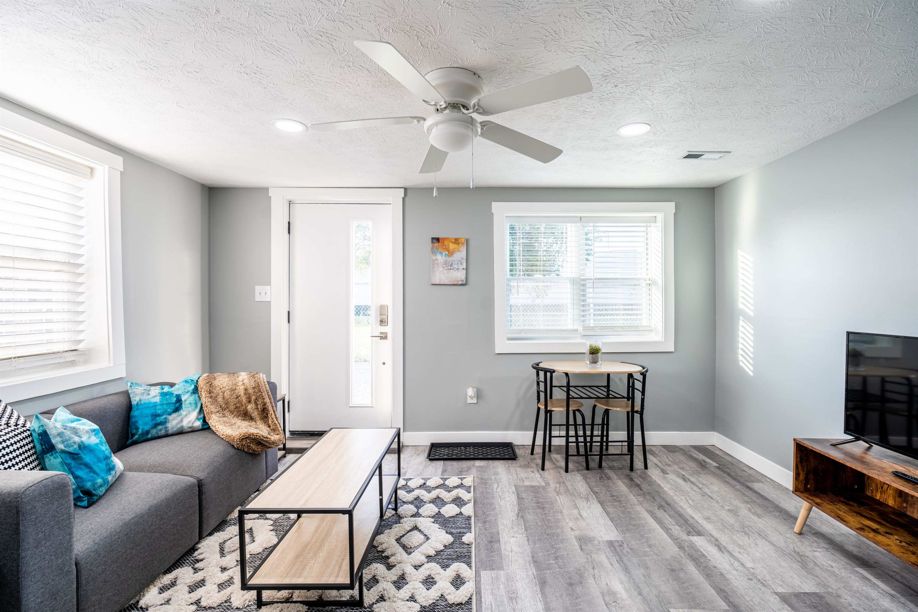 The living room features a gray sofa, a coffee table, a small dining table, and a wall-mounted television, all arranged around a light gray color scheme.