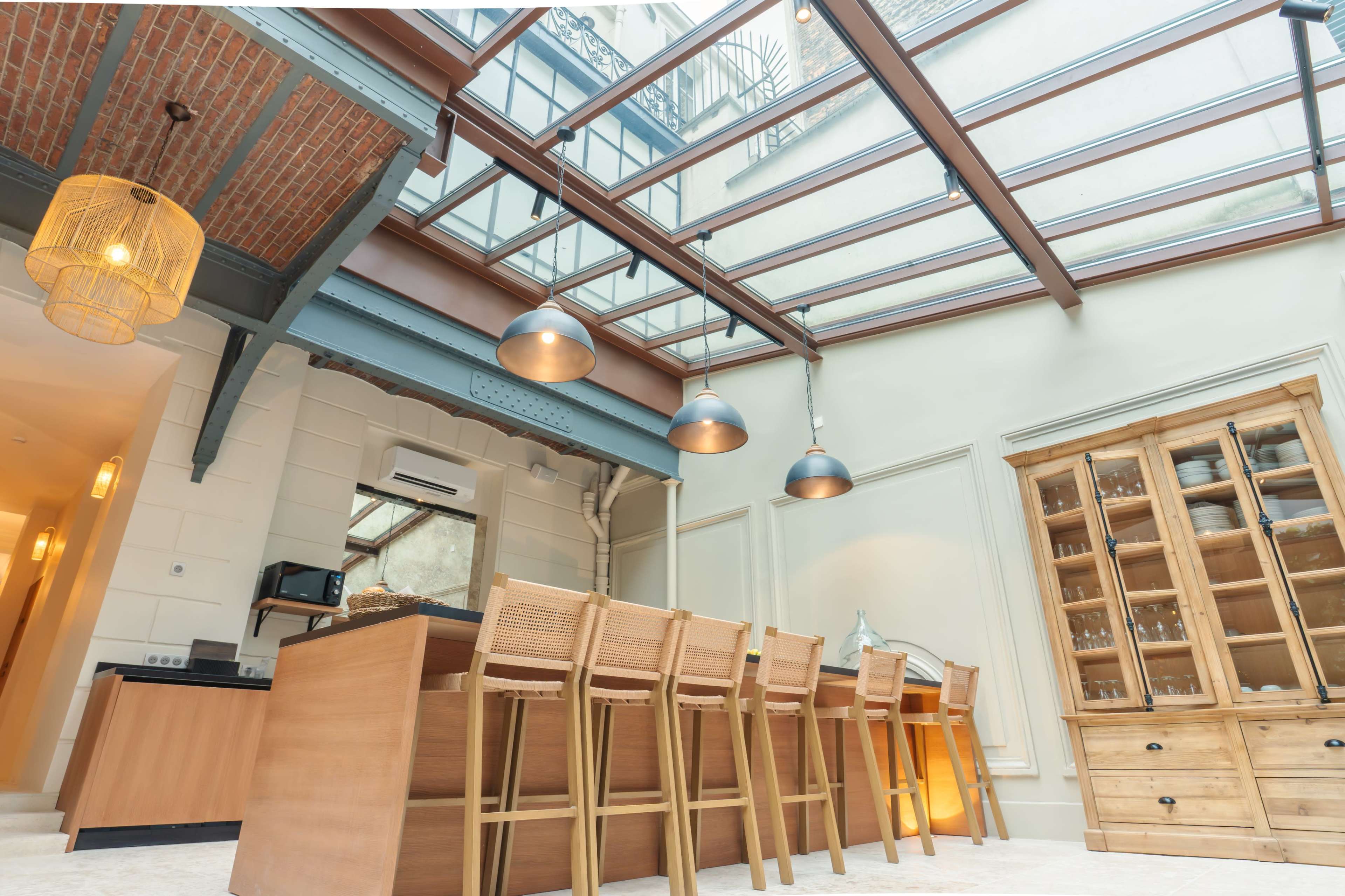 The image shows a modern kitchen with a glass ceiling, featuring wooden cabinets and a long dining table with high chairs.