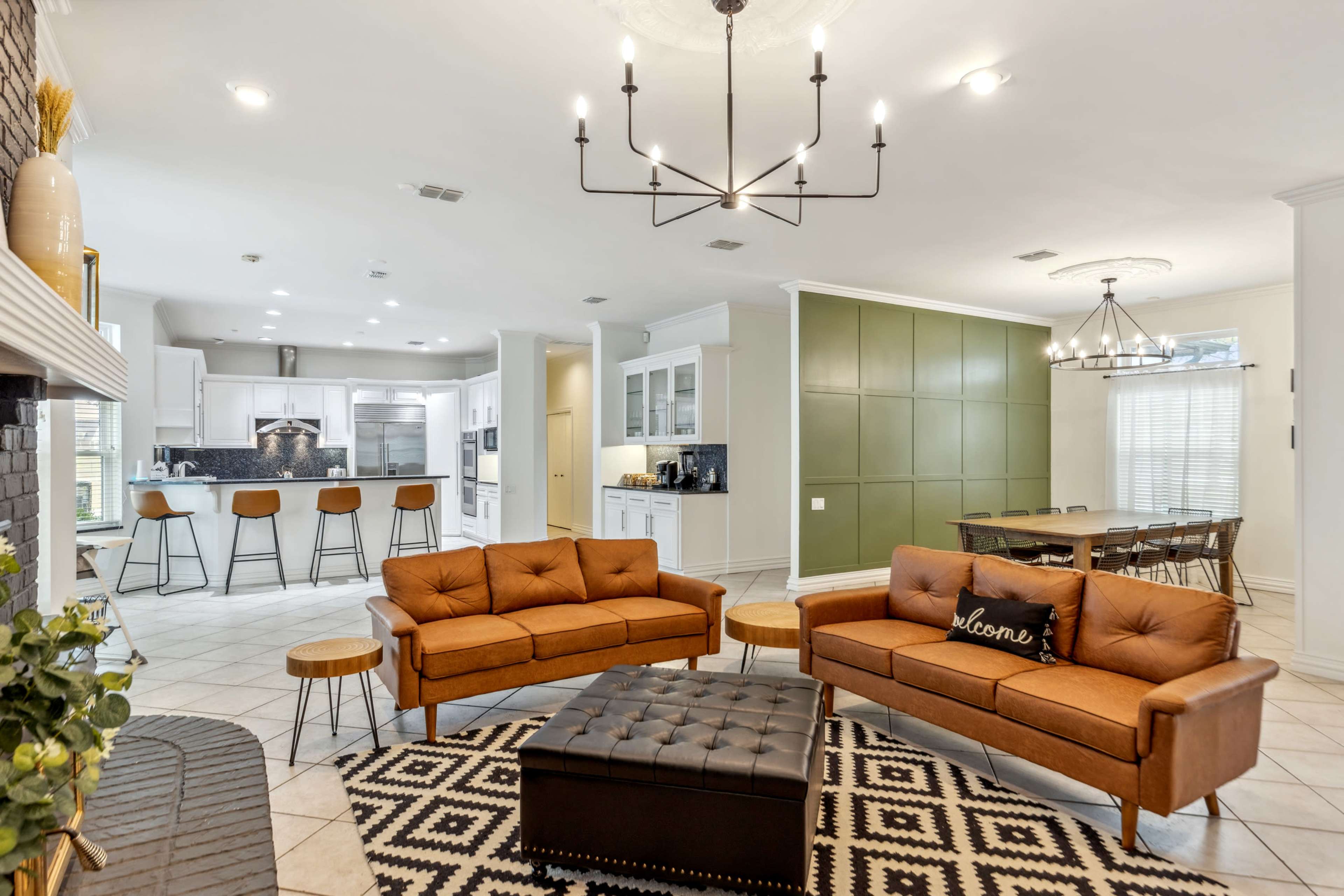 The image shows a modern, open-concept living area featuring two brown sofas, a coffee table, and a large kitchen with bar seating and green accent walls.