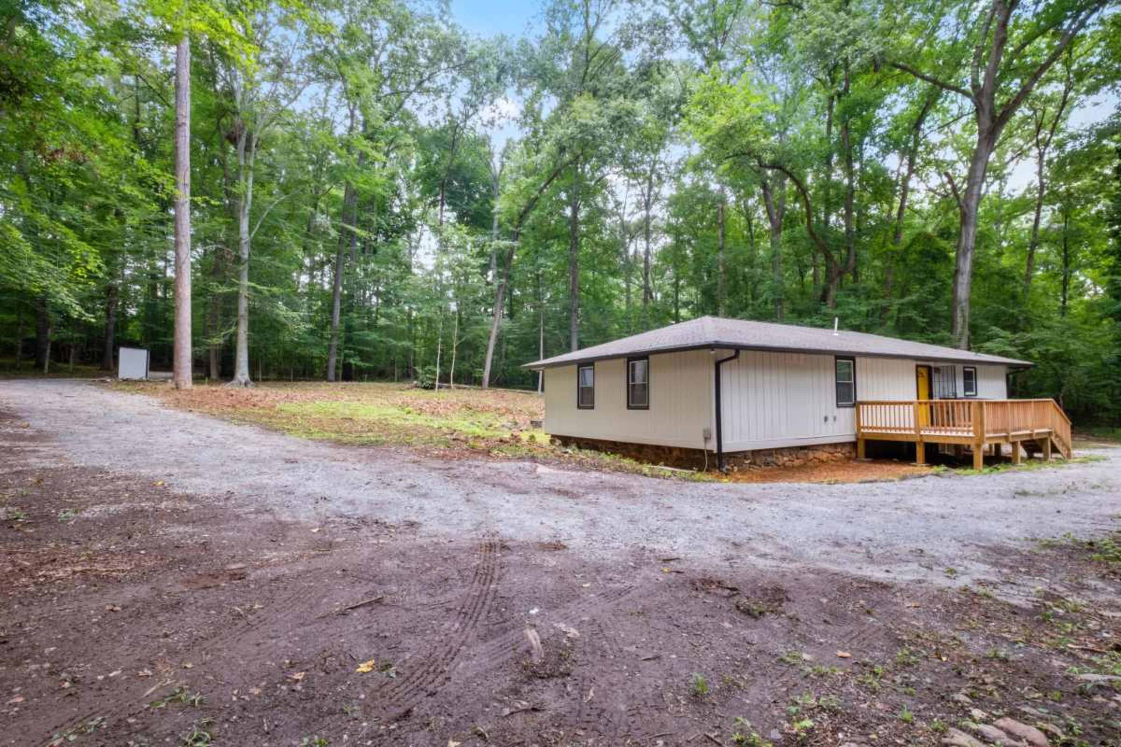 A single-story house with a wooden porch is positioned near a gravel driveway surrounded by dense trees.