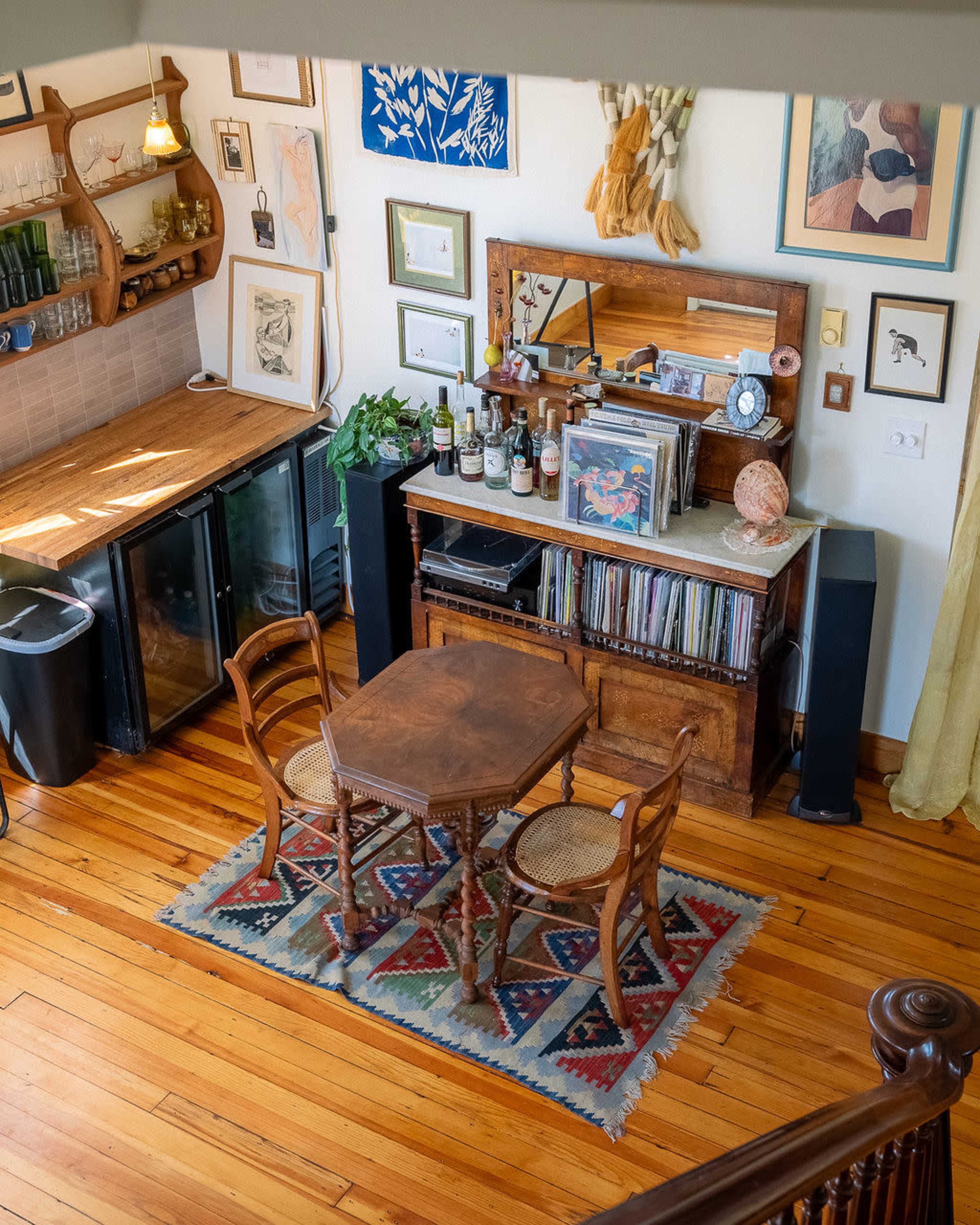 The image shows a cozy dining area with a wooden table and three chairs, surrounded by shelves displaying plants, artwork, and various decorative items.