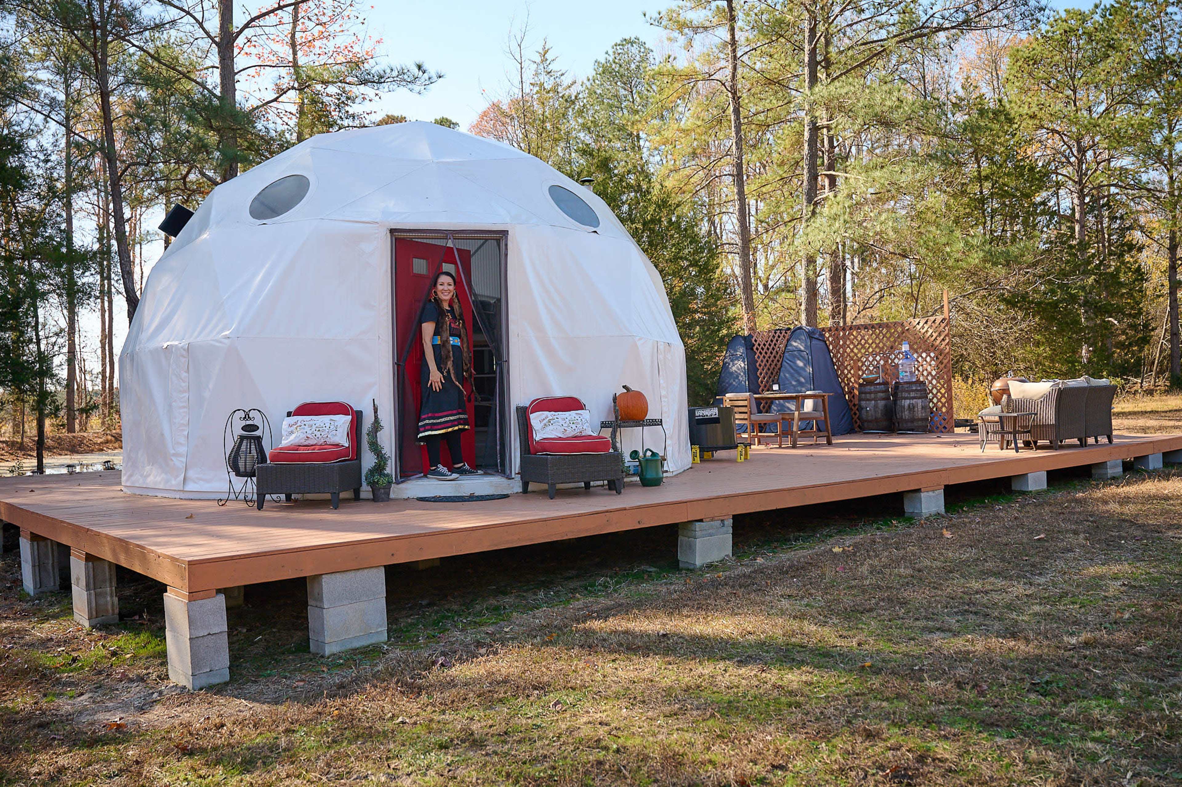 A woman stands in the doorway of a white geodesic dome home situated on a wooden deck surrounded by trees.