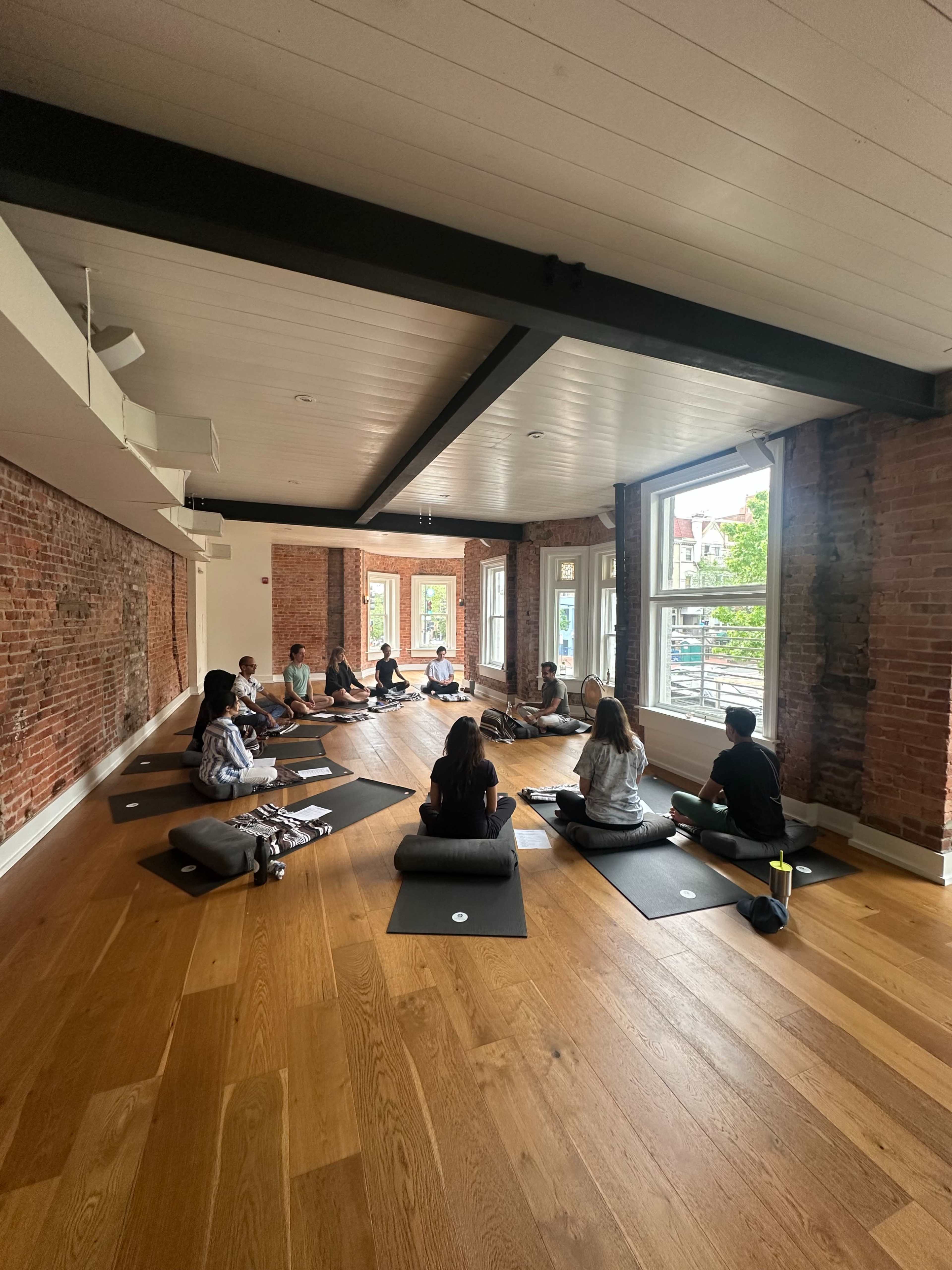 A group of individuals is seated on yoga mats in a spacious room with wooden floors and large windows, surrounded by brick walls.