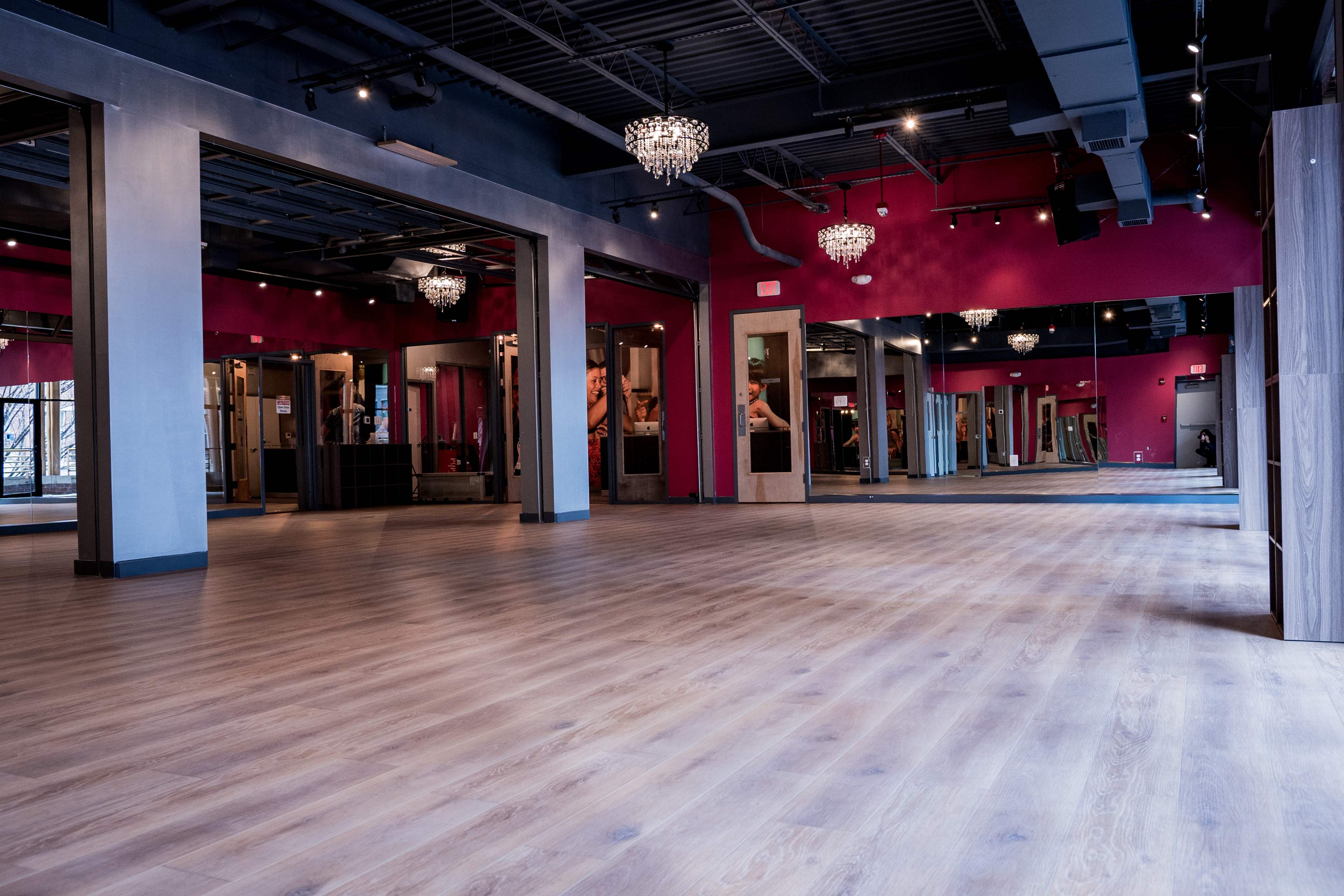 An empty fitness studio with wooden flooring, pink walls, and chandeliers reflecting overhead lighting.