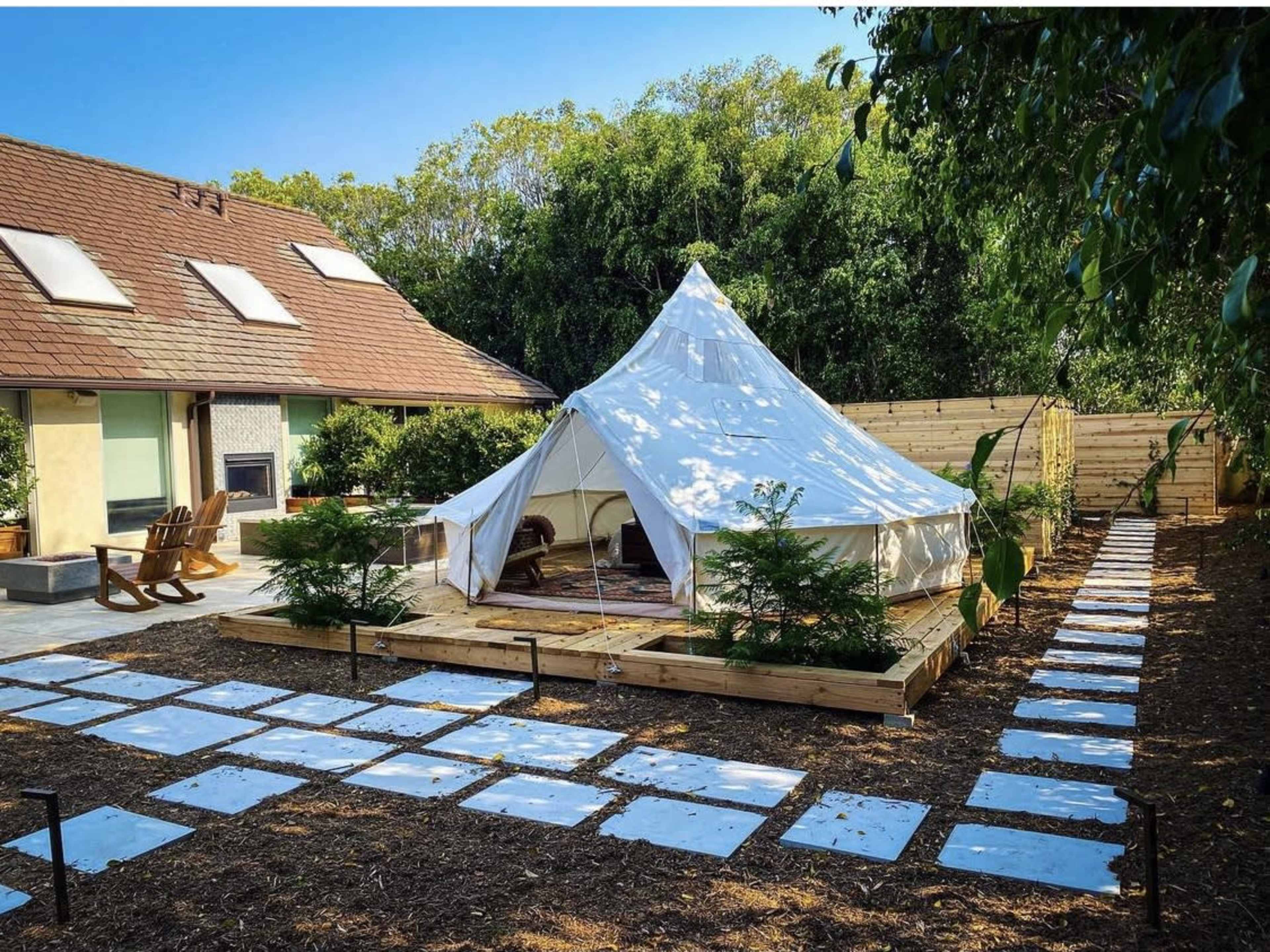 A large white bell tent is set on a wooden platform surrounded by a landscaped area with a stone path leading up to it.