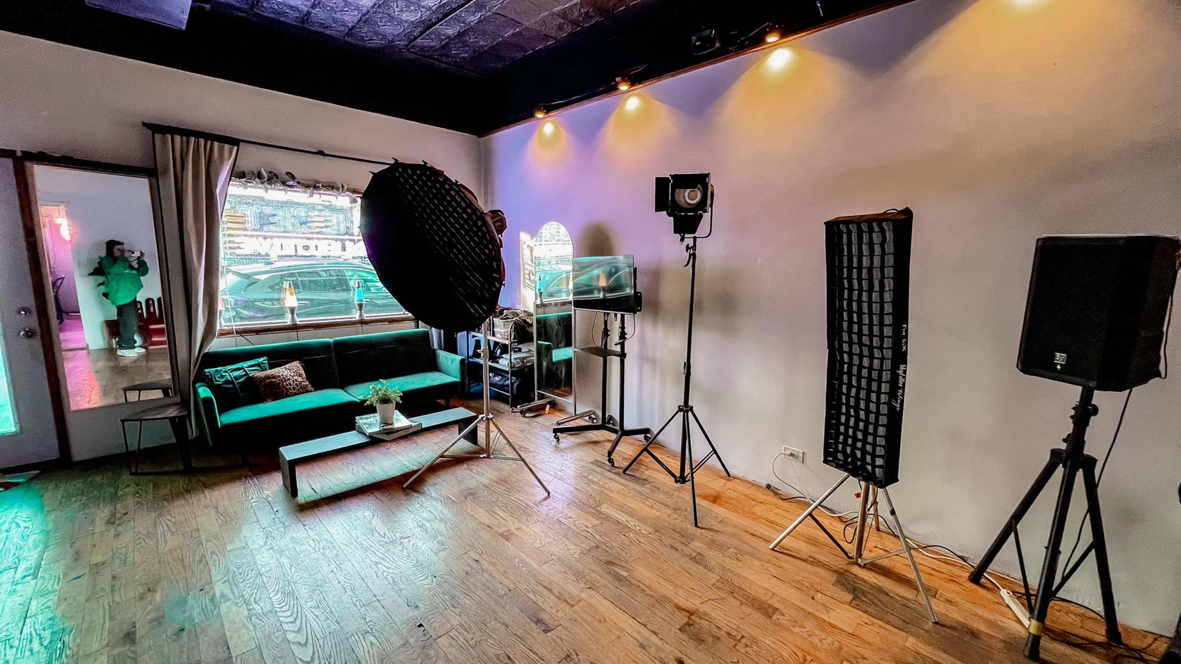 The image shows a room with a dark floor, a green couch, various lighting equipment, and a large umbrella softbox setup.