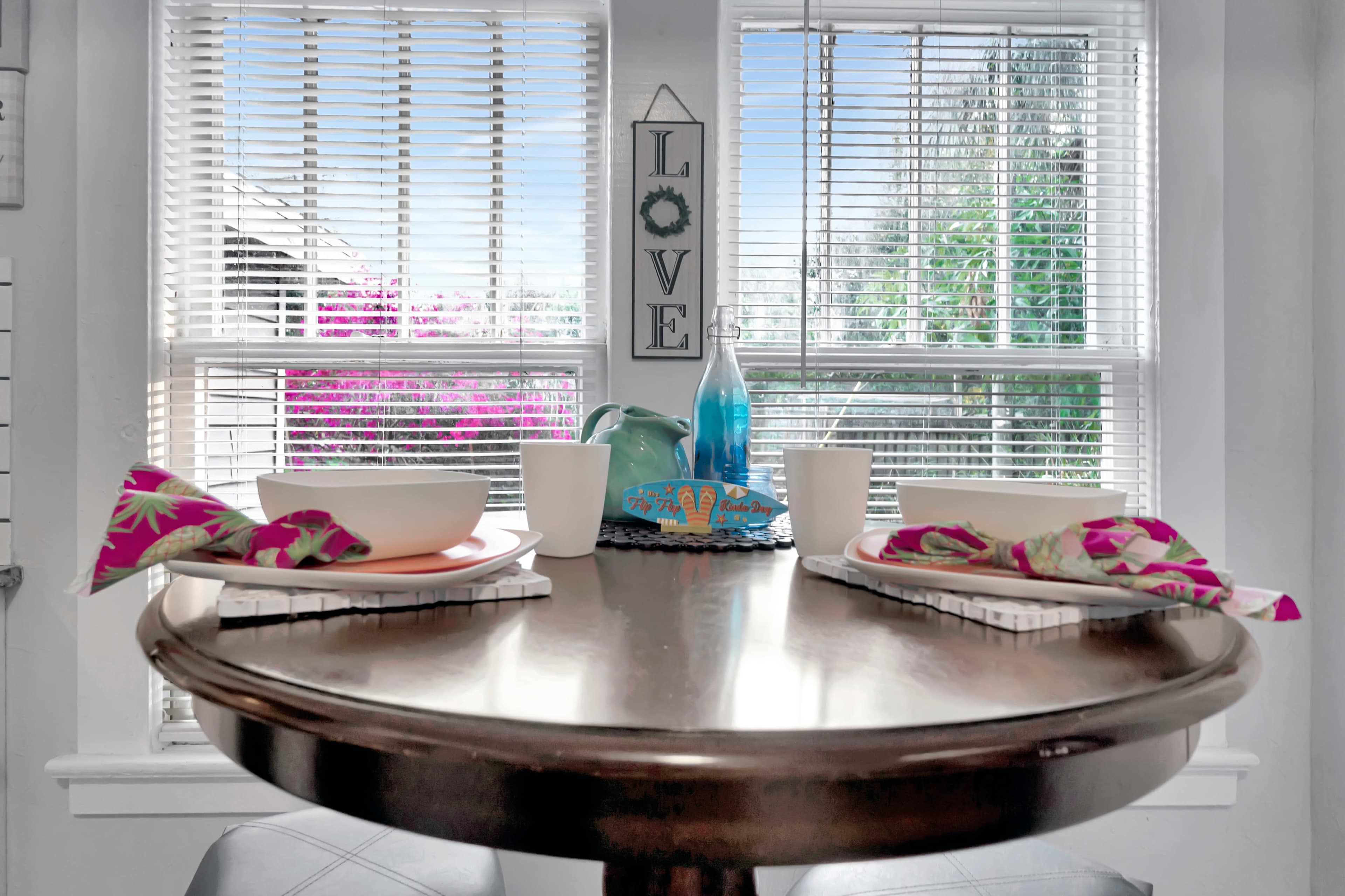 A wooden dining table is set for two with bowls, cups, and napkins, positioned near large windows adorned with blinds and overlooking outdoor plants.