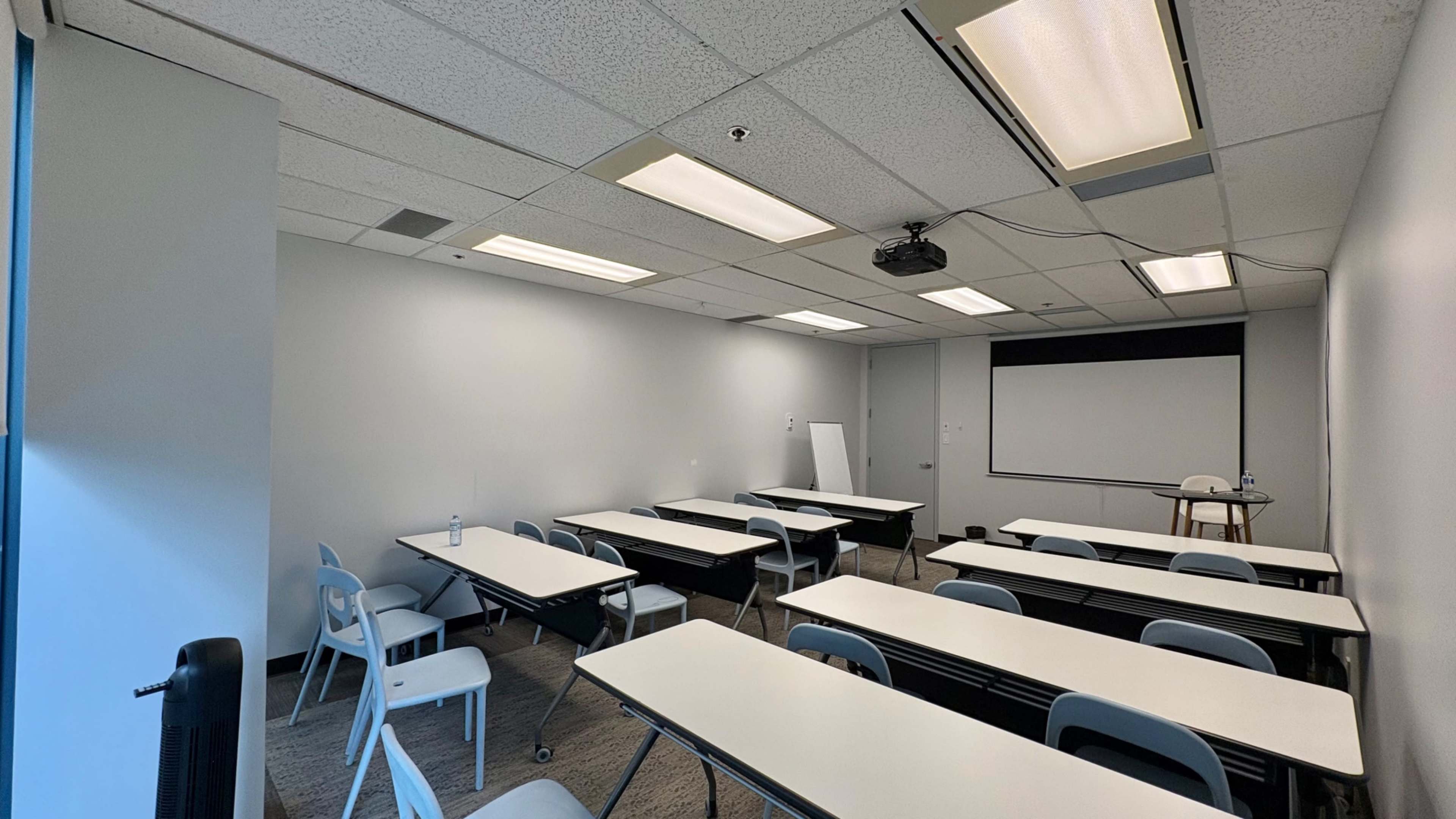 A spacious classroom features multiple tables arranged in rows, equipped with chairs and a projector screen at one end.
