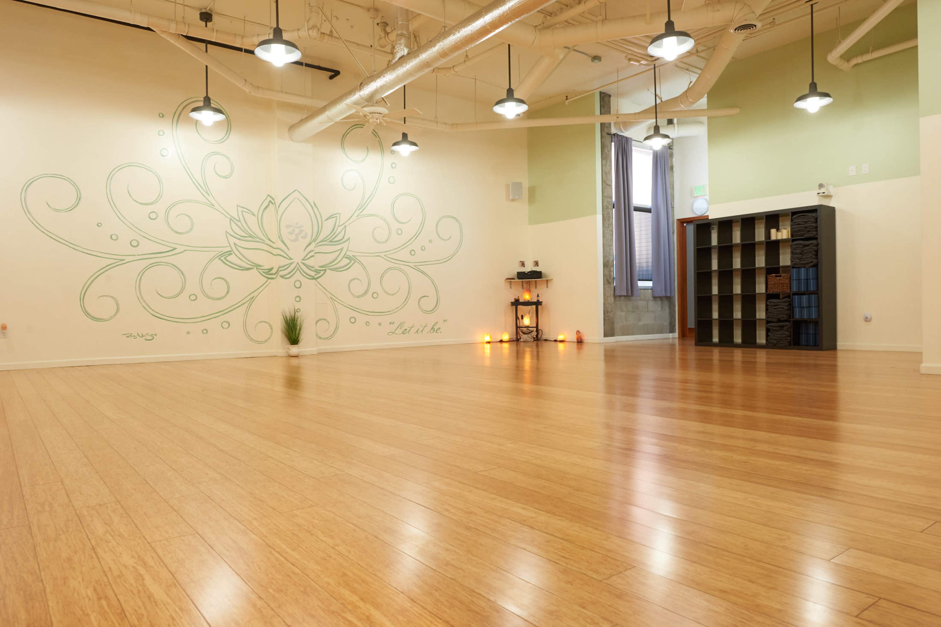 A spacious, well-lit yoga studio with bamboo flooring, a lotus flower mural on one wall, and shelves for storage on another.