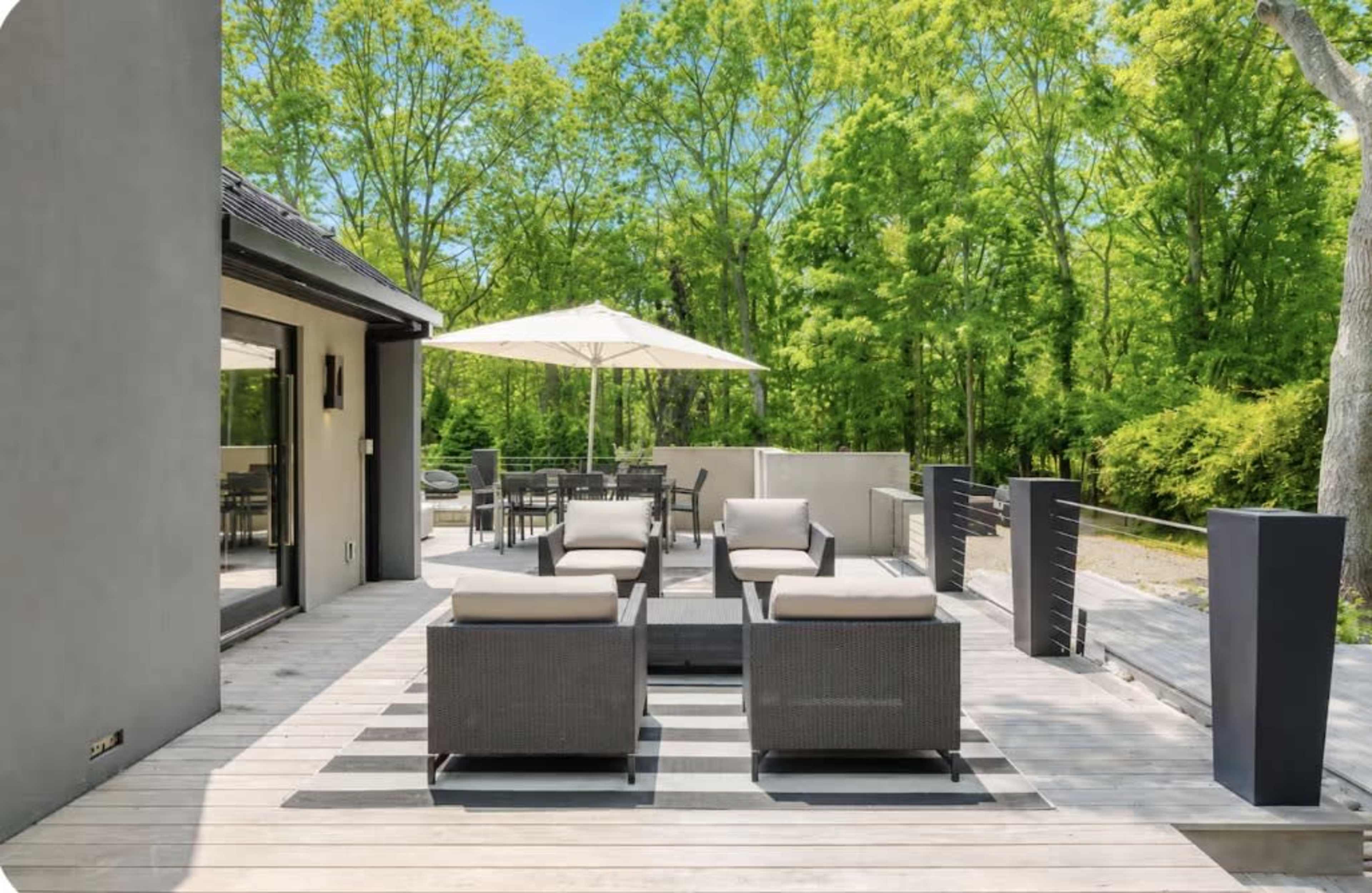 The image shows a modern outdoor patio furnished with seating and a dining area, surrounded by lush green trees.