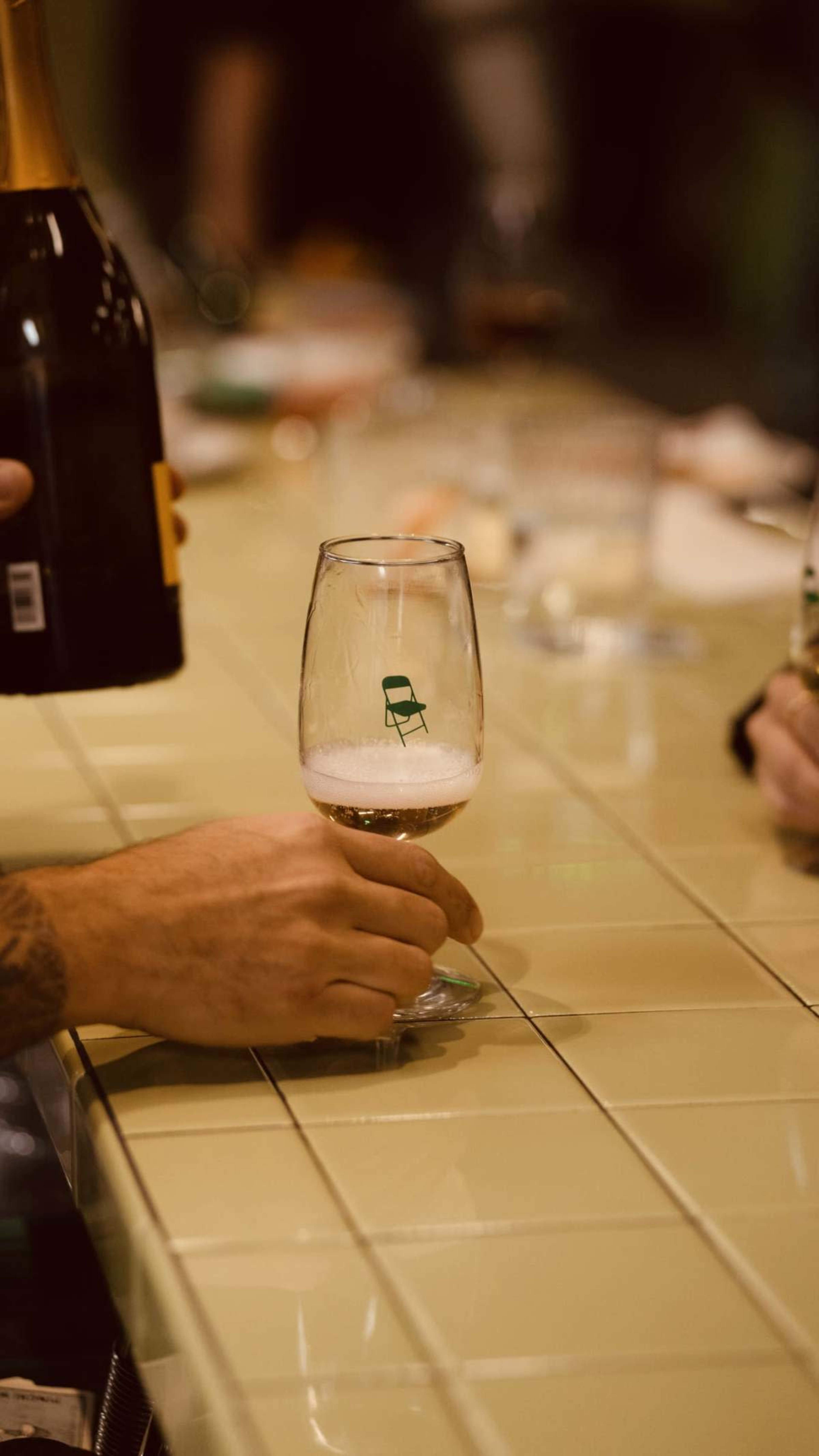 A person holds a glass with a green chair logo while another bottle is being poured nearby on a tiled bar surface.