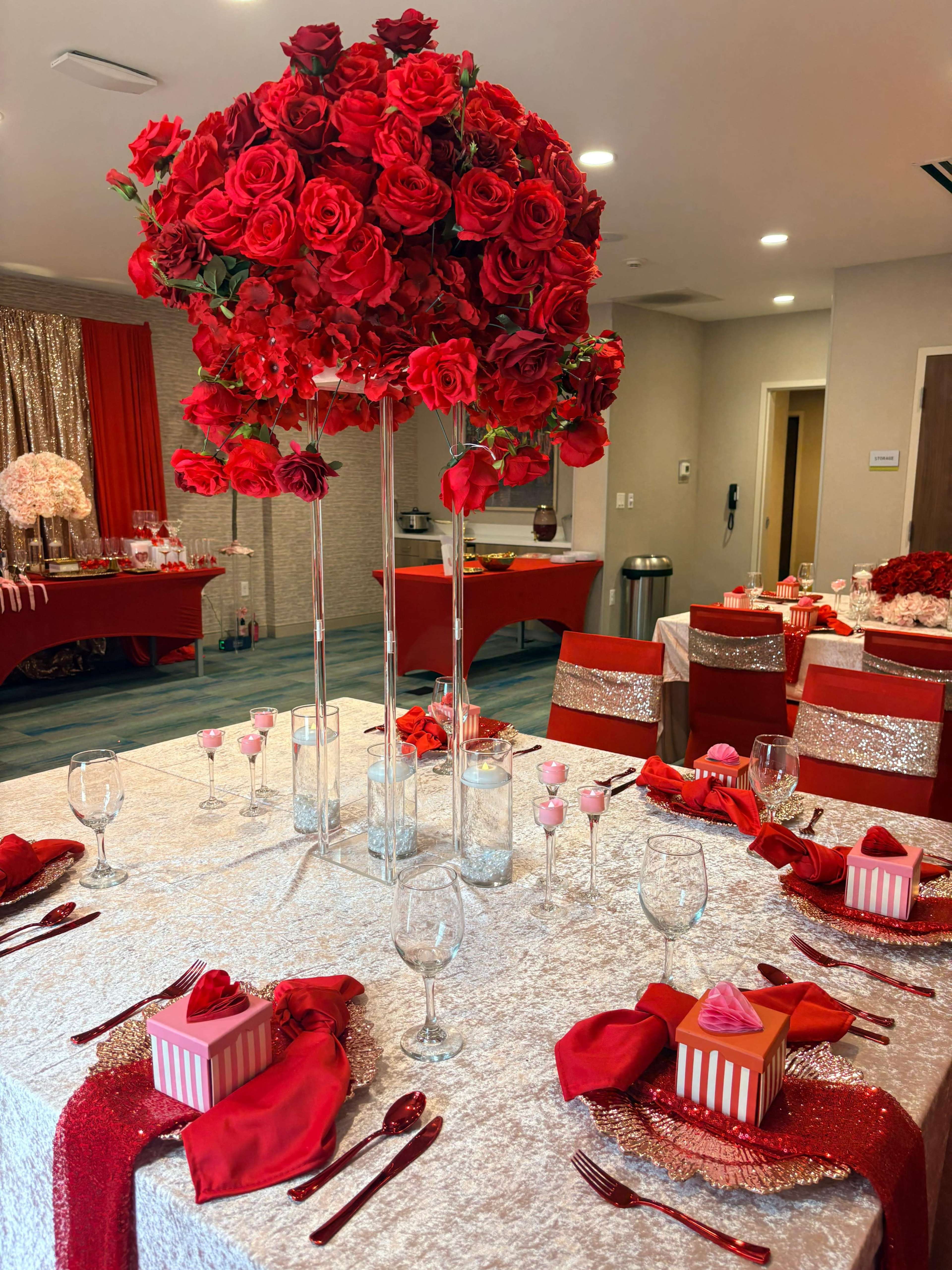 A dining table is elegantly arranged with a silver tablecloth, tall floral centerpieces of red roses, and decorative place settings featuring glassware and small gift boxes.