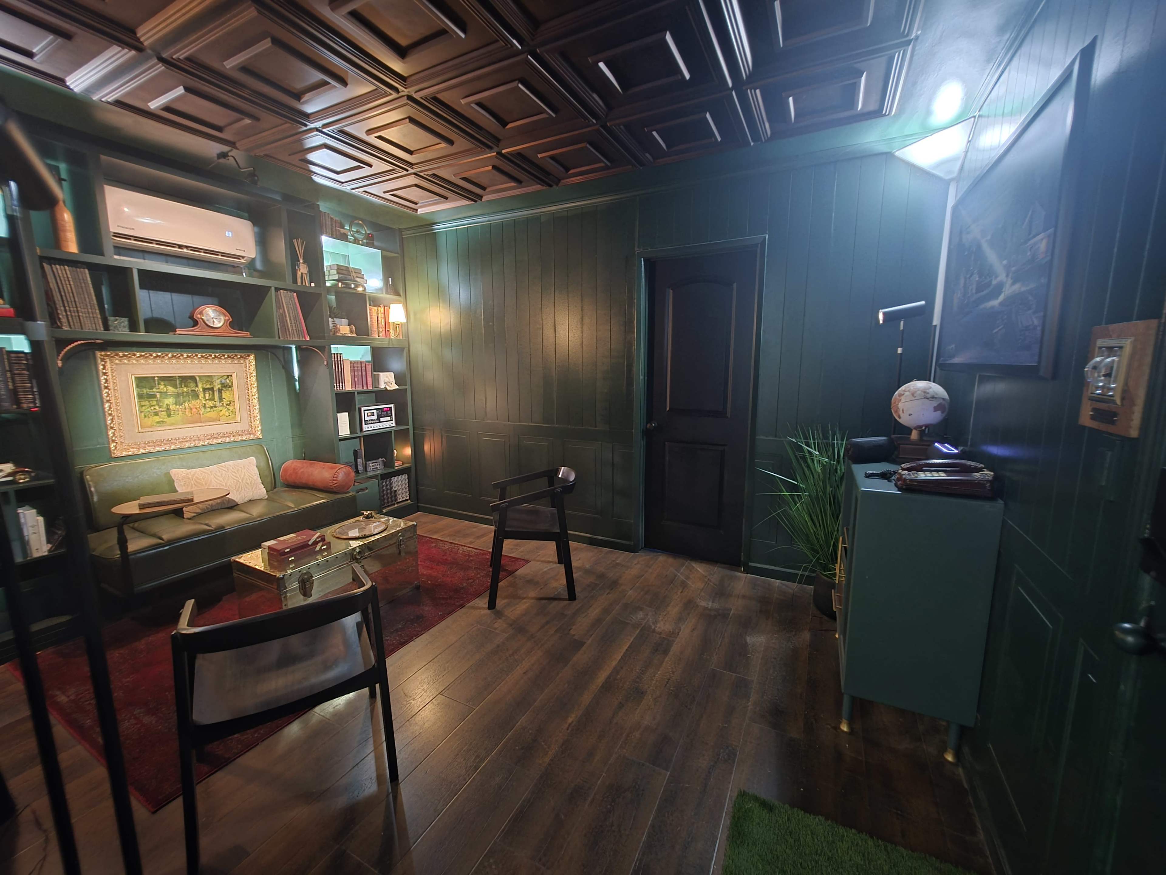 The image shows a cozy room with dark green walls, a wooden coffered ceiling, a couch, a bookshelf, and decorative items, along with a door and a potted plant.