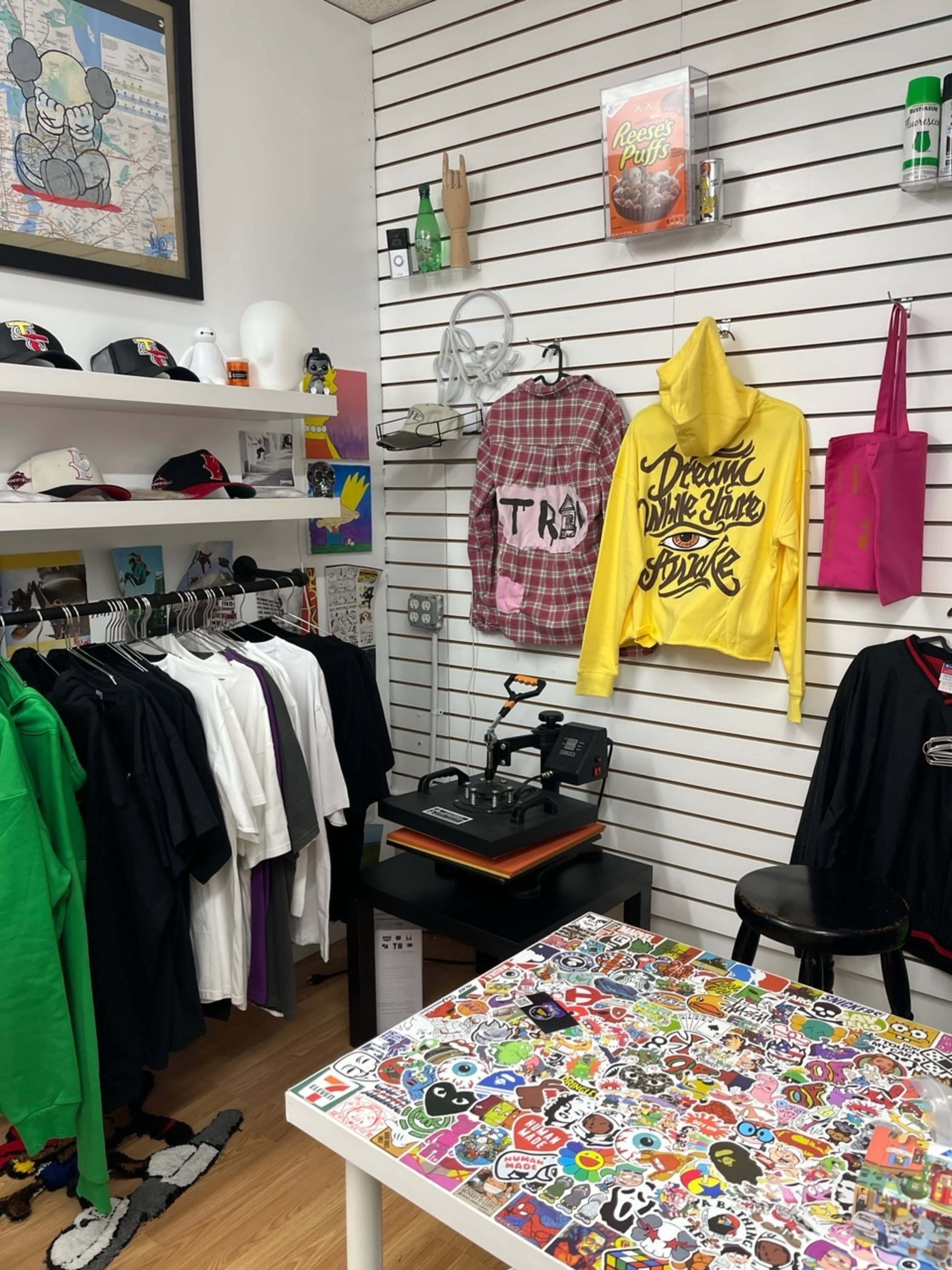 The image shows a small retail space featuring a variety of clothing items, including hoodies and t-shirts, hanging on white walls alongside a colorful table adorned with numerous stickers.