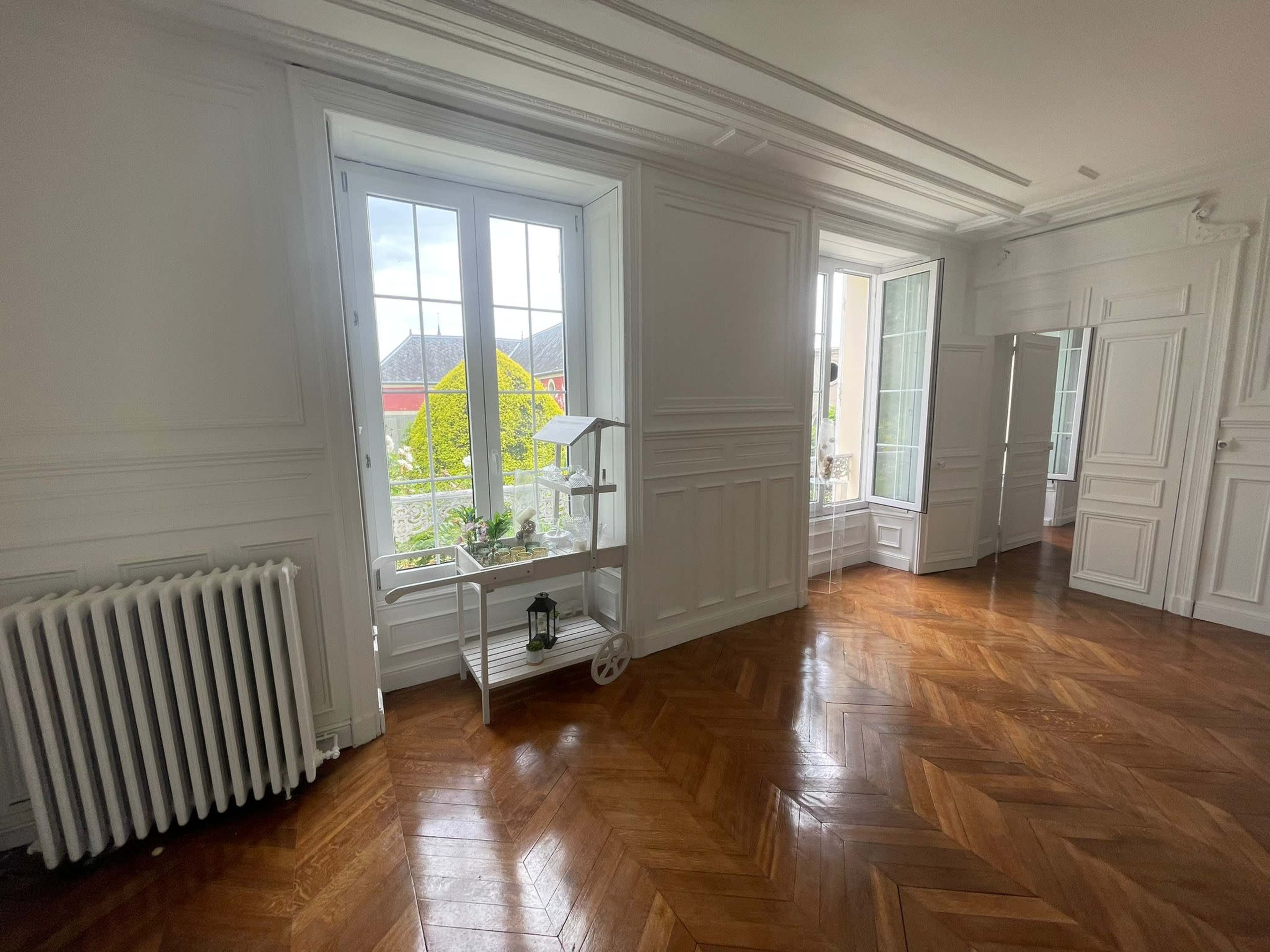 A brightly lit room with large windows, a wooden herringbone floor, and a white radiator.