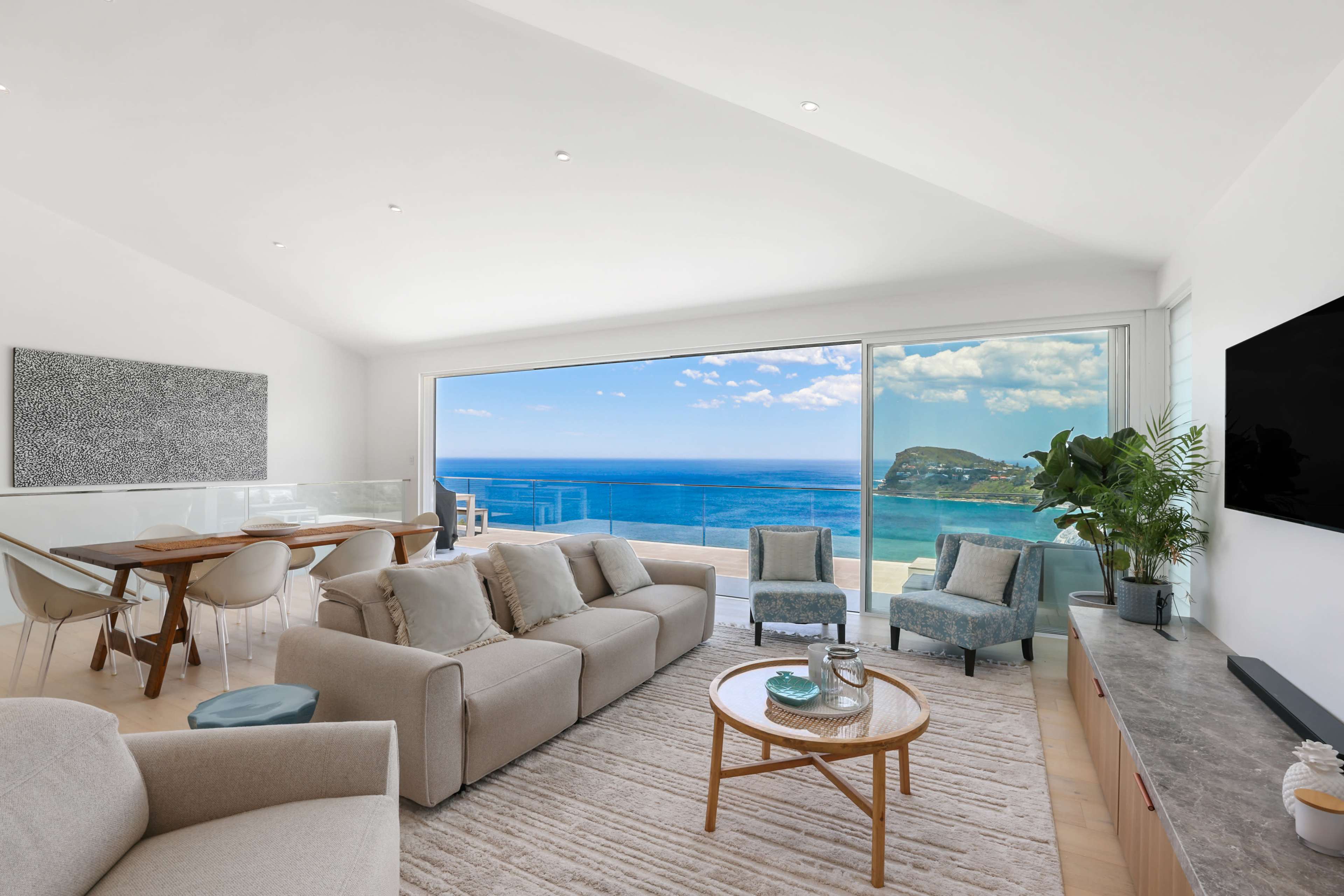 A modern living room features a large window with a view of the ocean, furnished with a beige sofa, a wooden coffee table, and a dining area in the background.