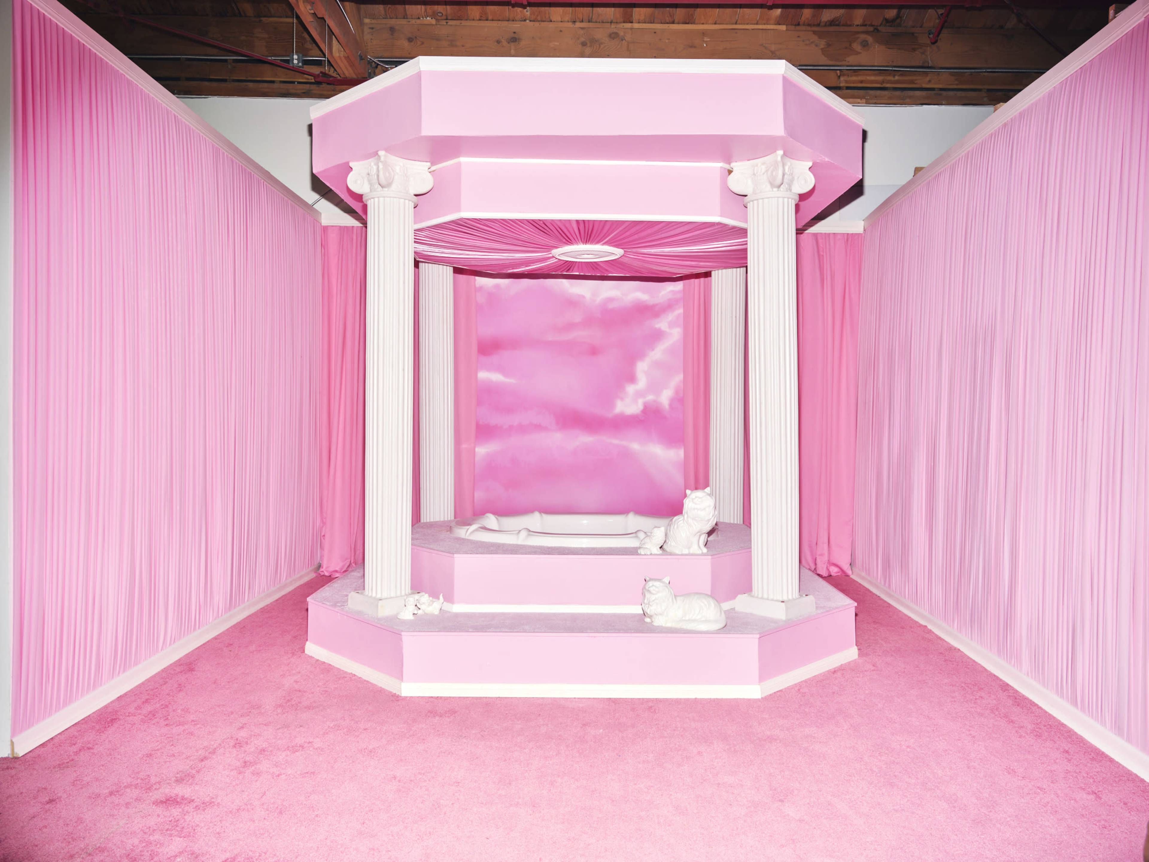 The image shows a pink room with a raised pedestal featuring a bath surrounded by four white columns and draped pink curtains.