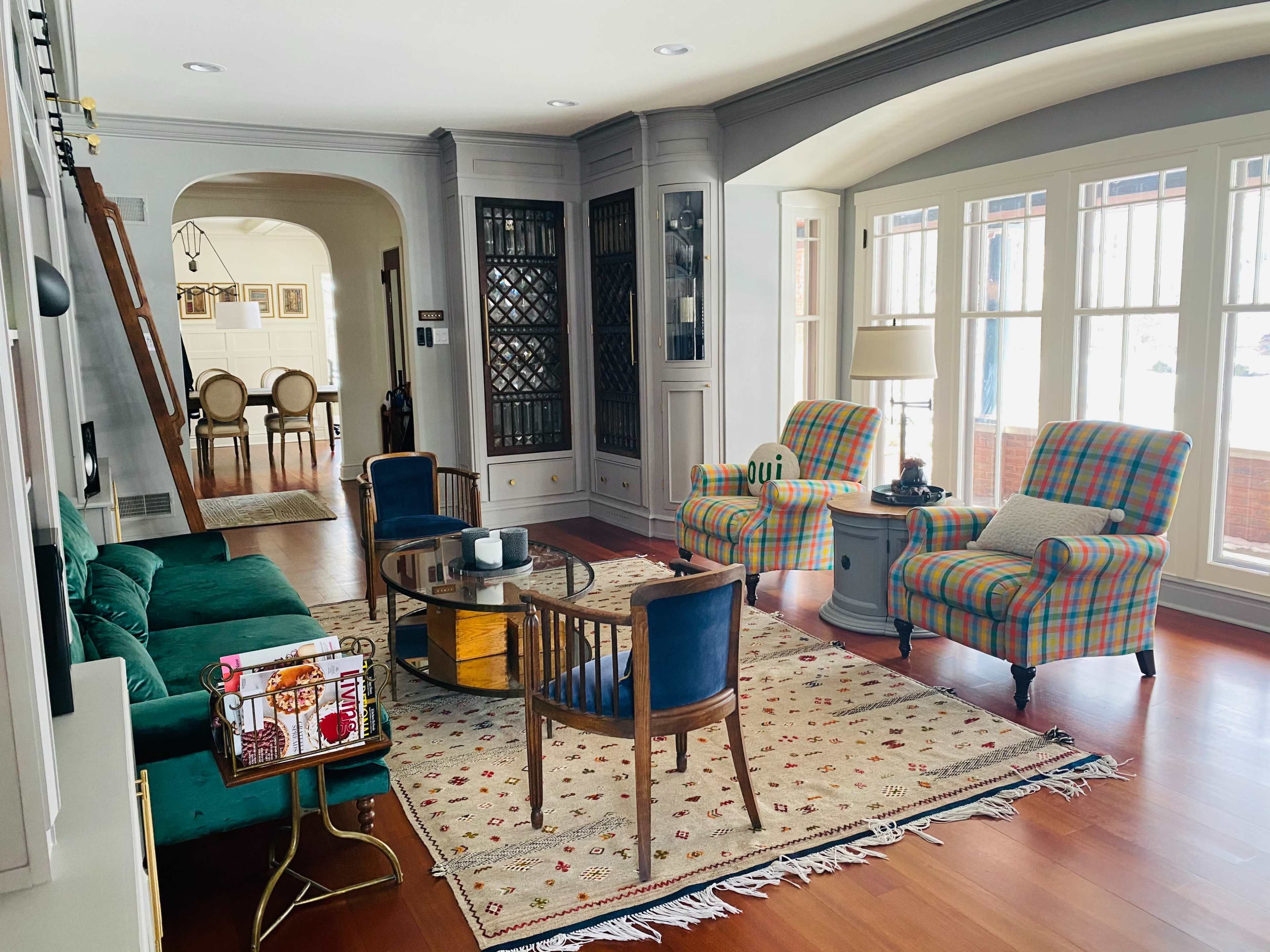 A cozy living room features two patterned armchairs, a green sofa, a round coffee table, and large windows allowing ample natural light.