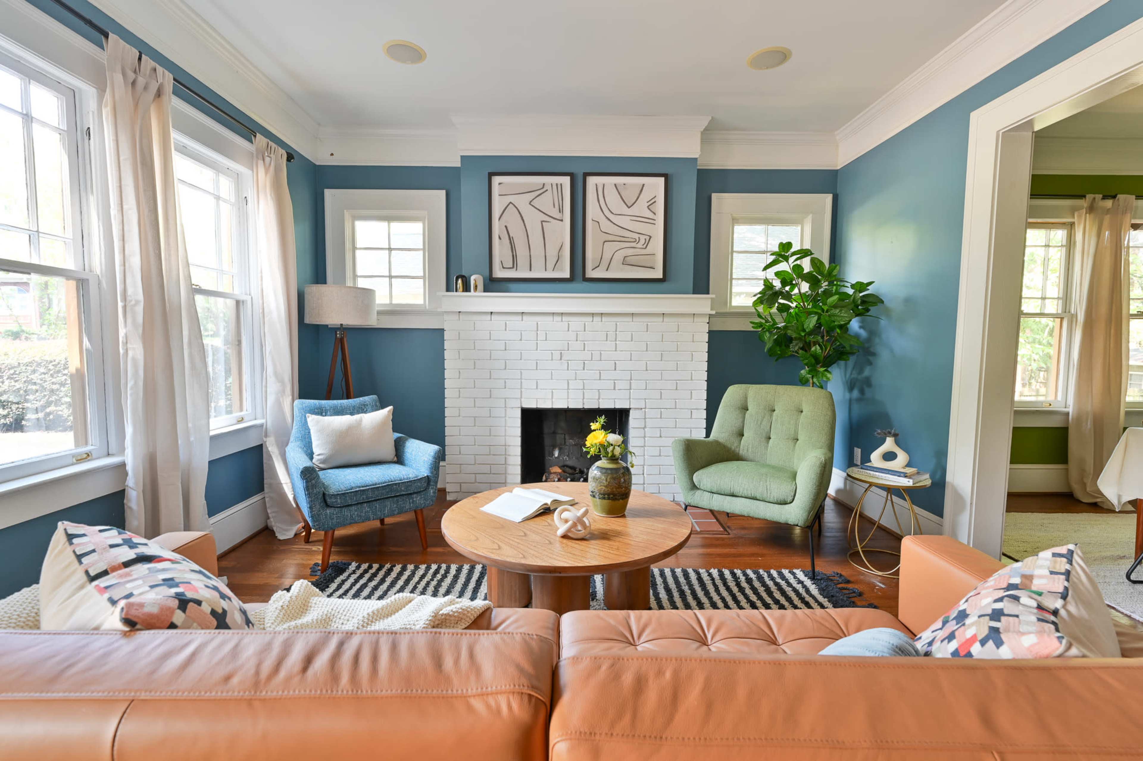 A living room featuring a blue wall, a white brick fireplace, a round wooden coffee table, and two distinct seating areas with a light brown sofa and chairs in blue and green tones.
