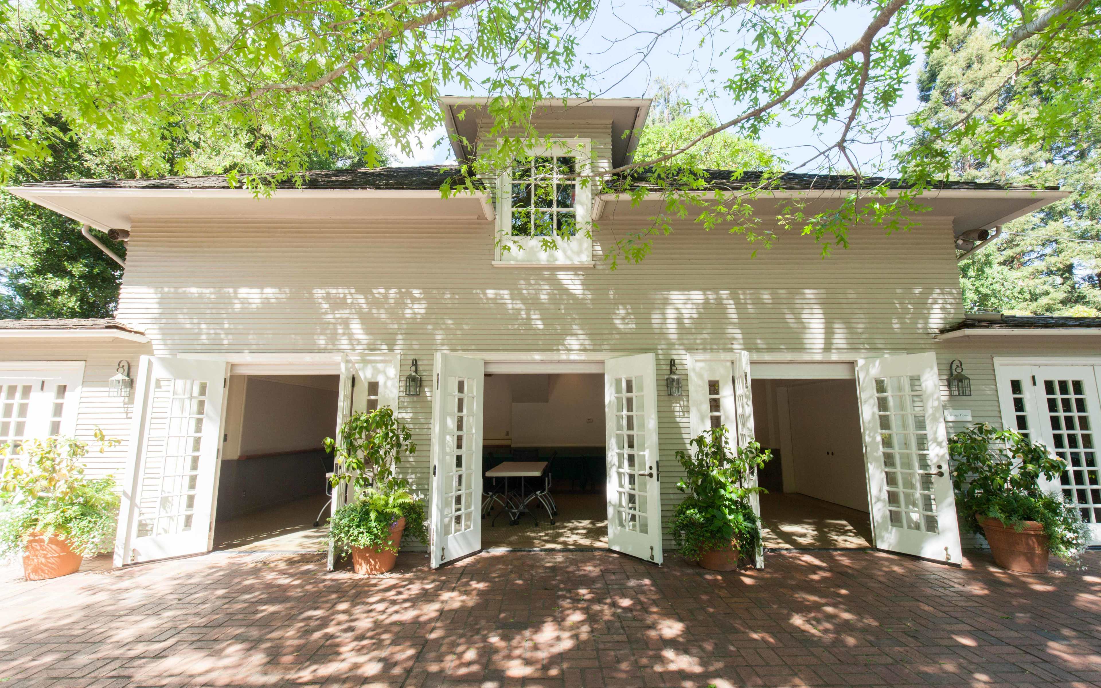 A light-colored building with multiple open French doors and potted plants on a brick patio, framed by leafy trees.
