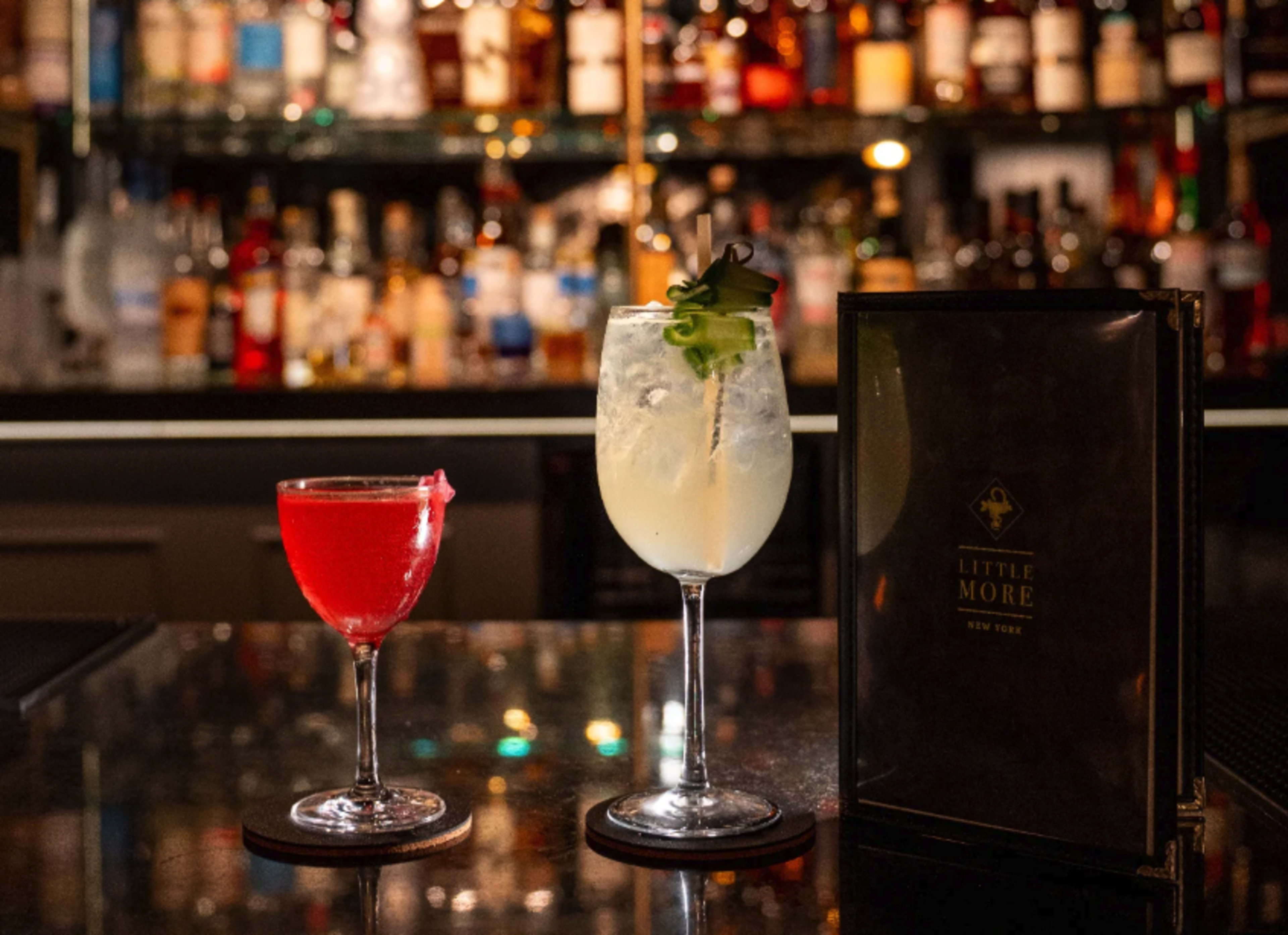 Two cocktails are displayed on a bar top, with a vibrant red drink in a coupe glass and a pale yellow drink in a large glass, accompanied by a menu in a black cover.