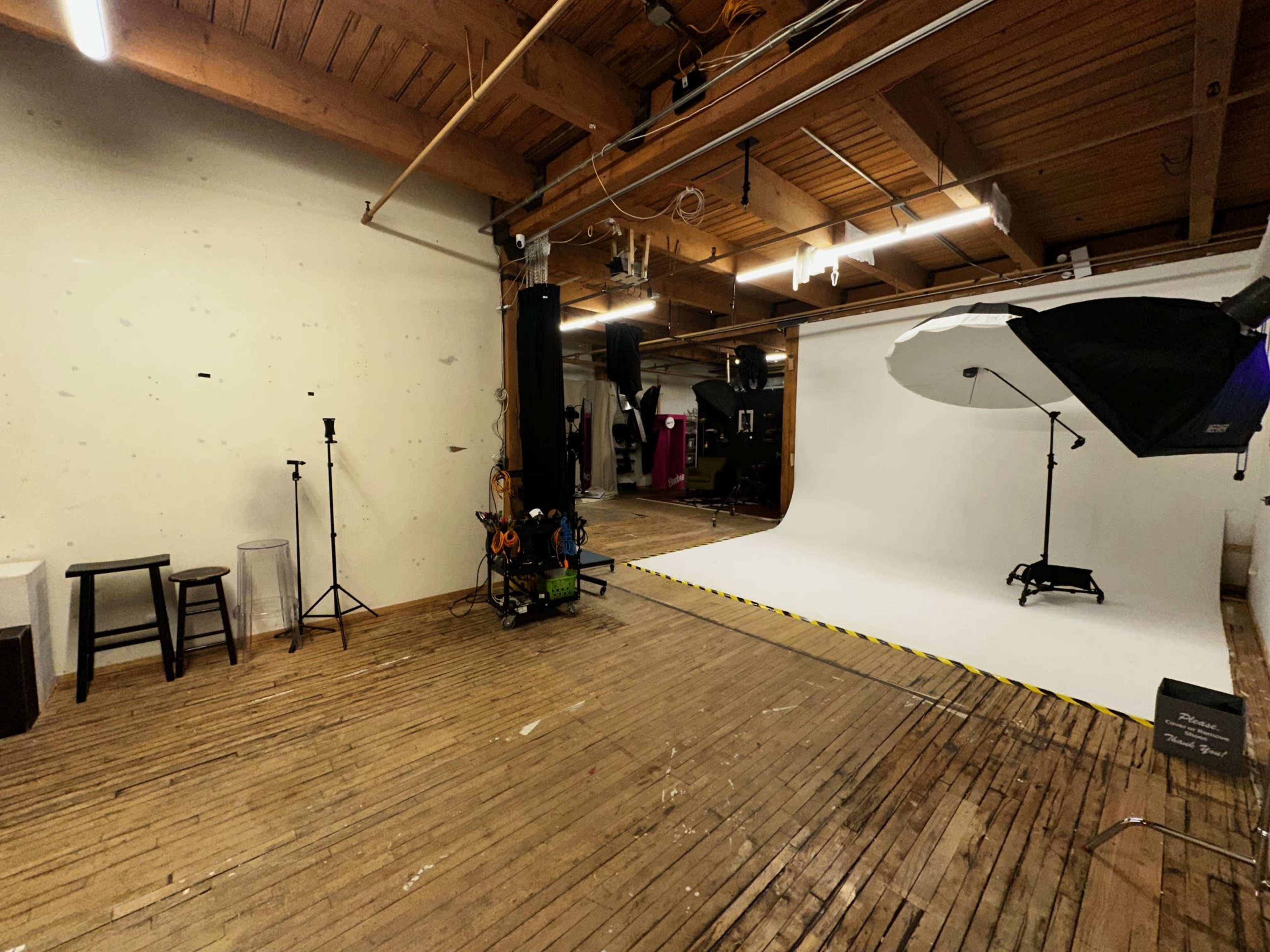 The image shows a photography studio featuring a large white backdrop, lighting equipment, and wooden flooring, with some furniture positioned in the corner.