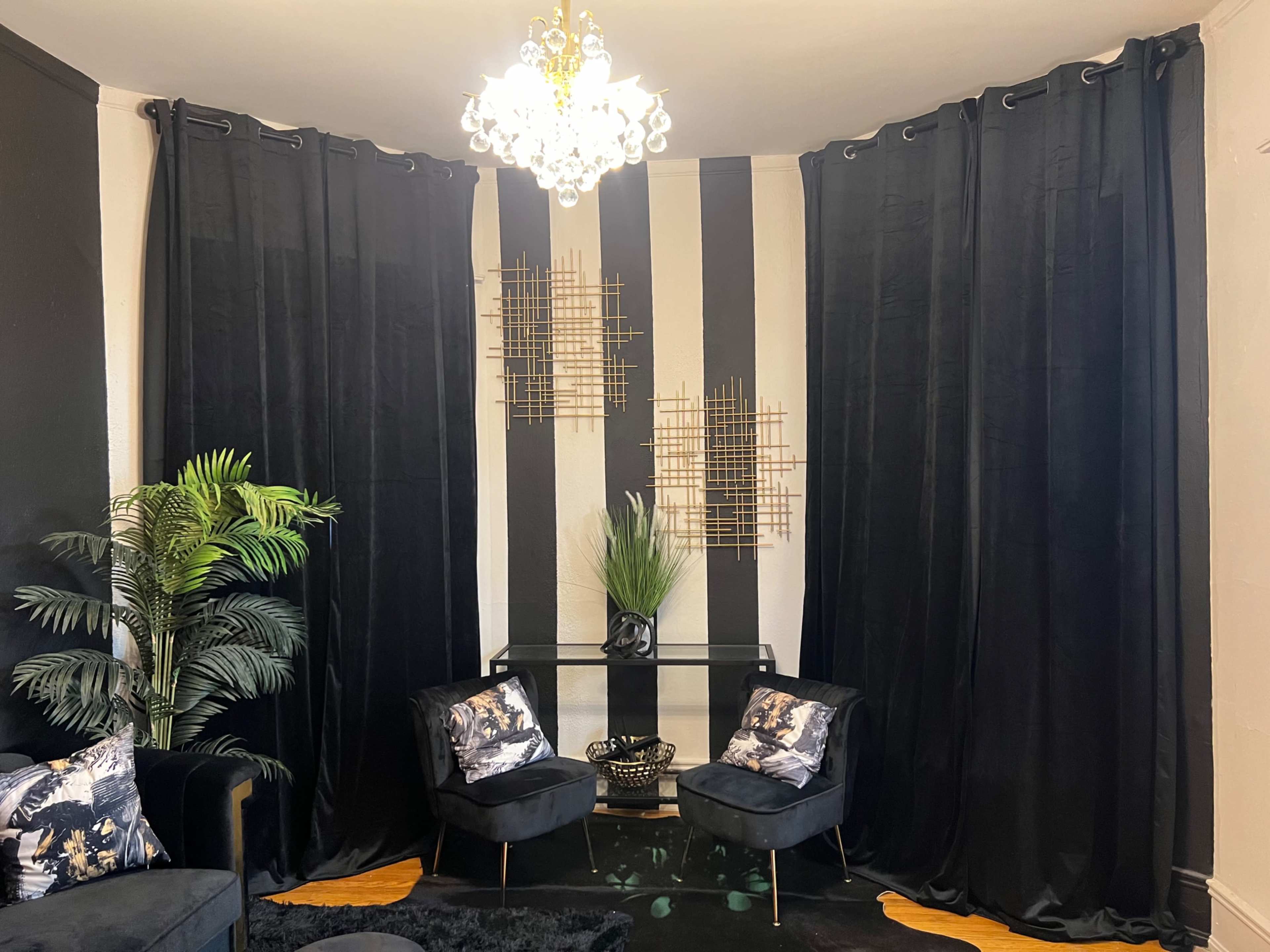 The image depicts a room with black and white striped walls, two black curtains, a glass table, two chairs with decorative cushions, and a chandelier overhead.
