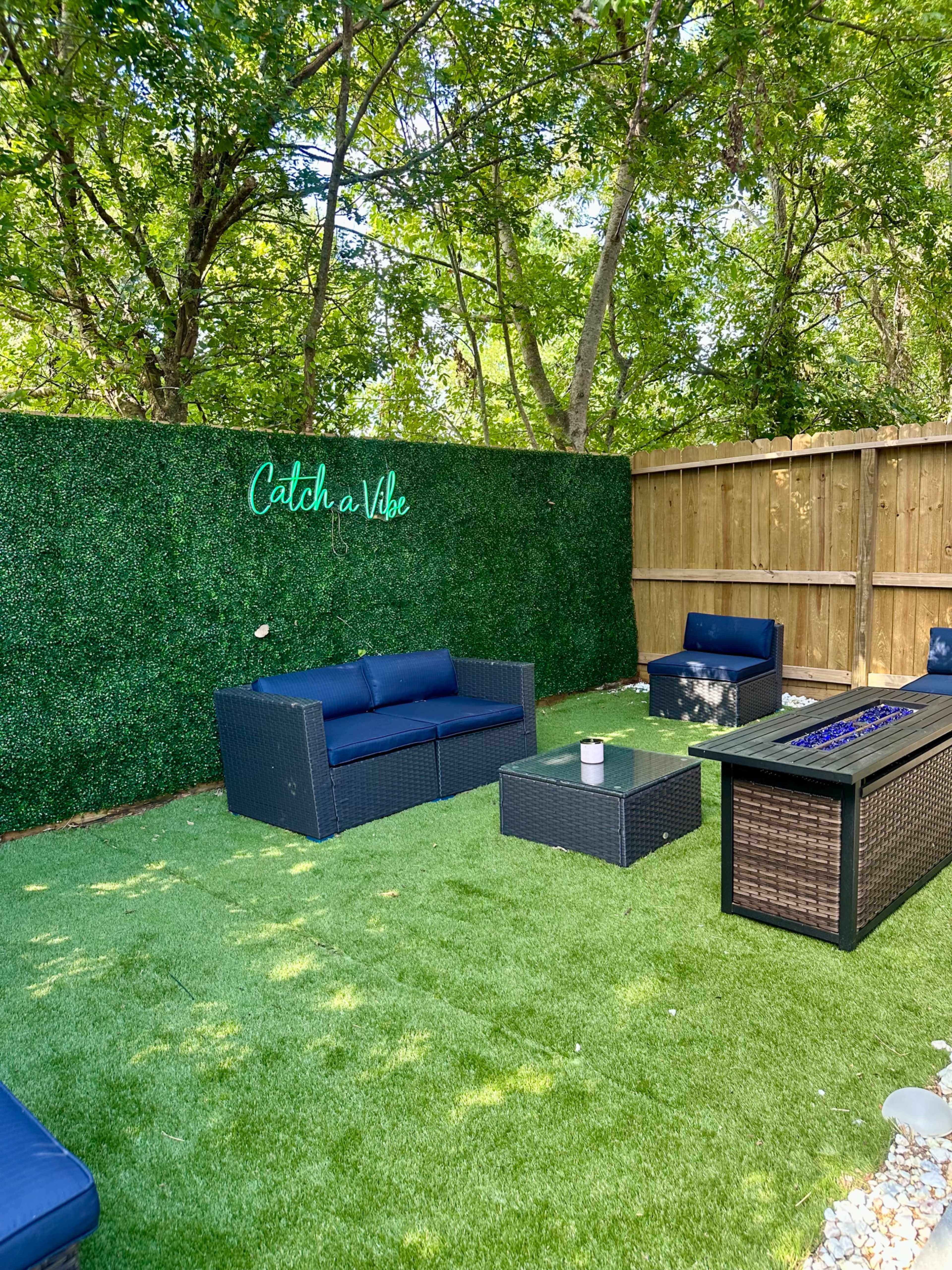 A landscaped outdoor space with synthetic grass, seating couches, a coffee table, and a fire pit against a green wall.