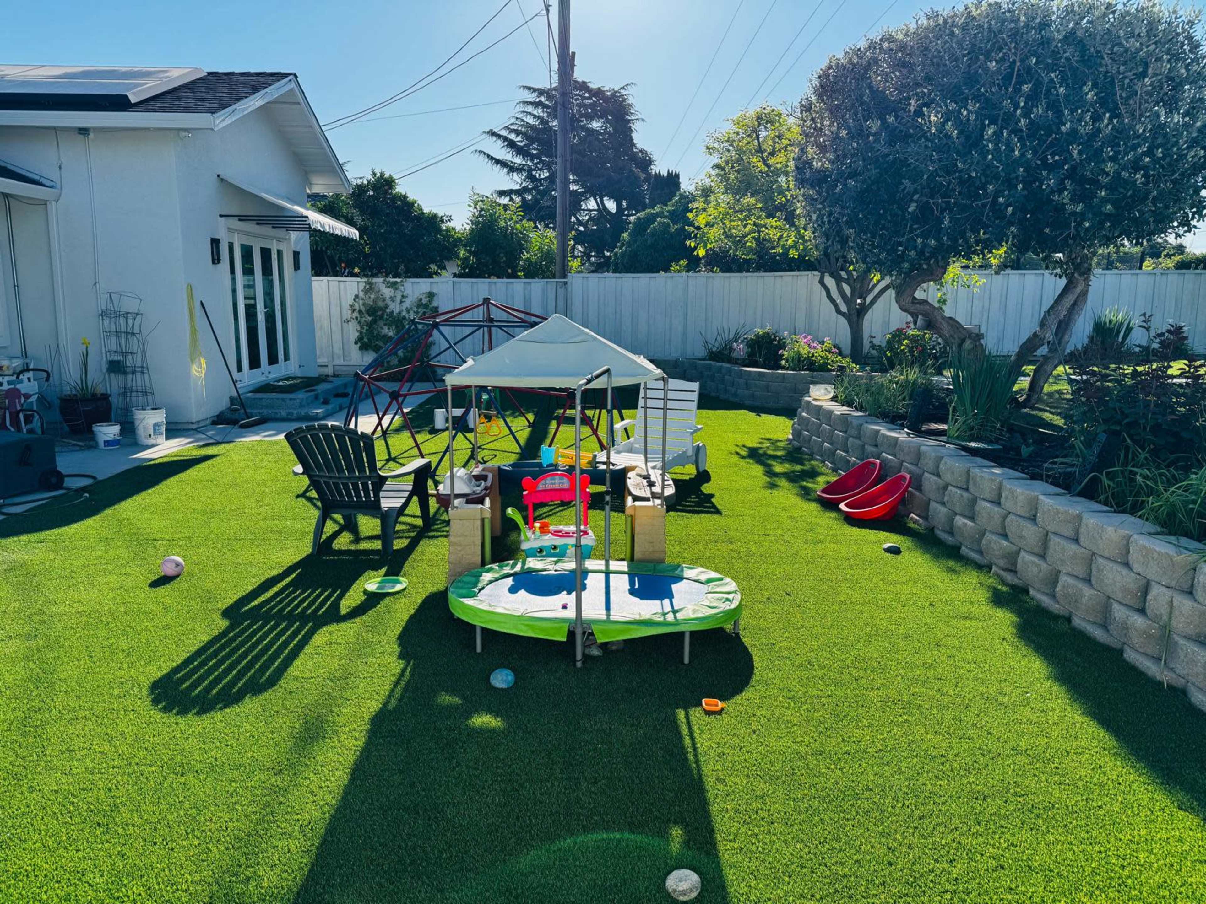 The image shows a backyard featuring a trampoline, a play structure, garden areas, and seating on artificial grass under clear skies.