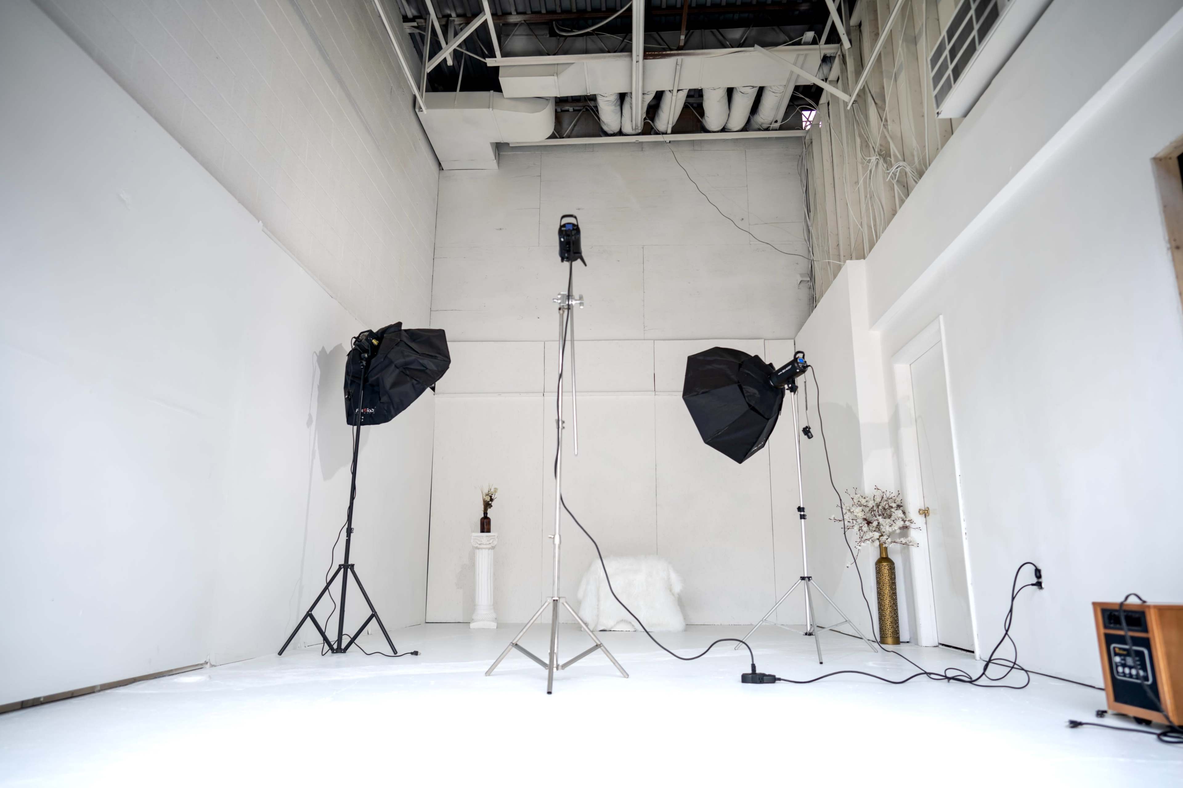 A photography studio with two softbox lights on tripods, a microphone on a stand, and minimal decor, all set against white walls and a white floor.