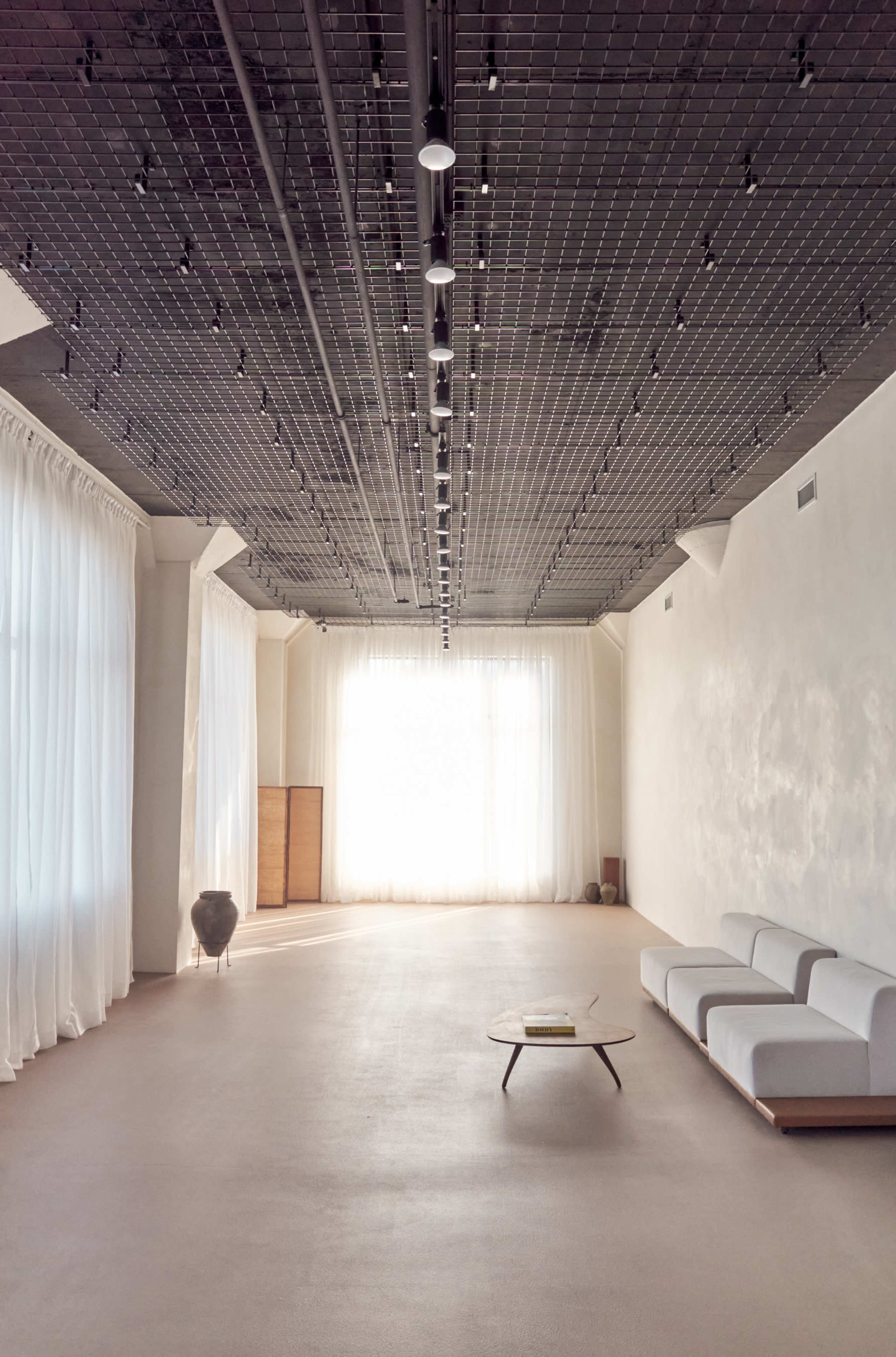 The image shows a spacious, minimalist interior with a light-colored floor, large windows covered by sheer curtains, and a simple sofa and coffee table.