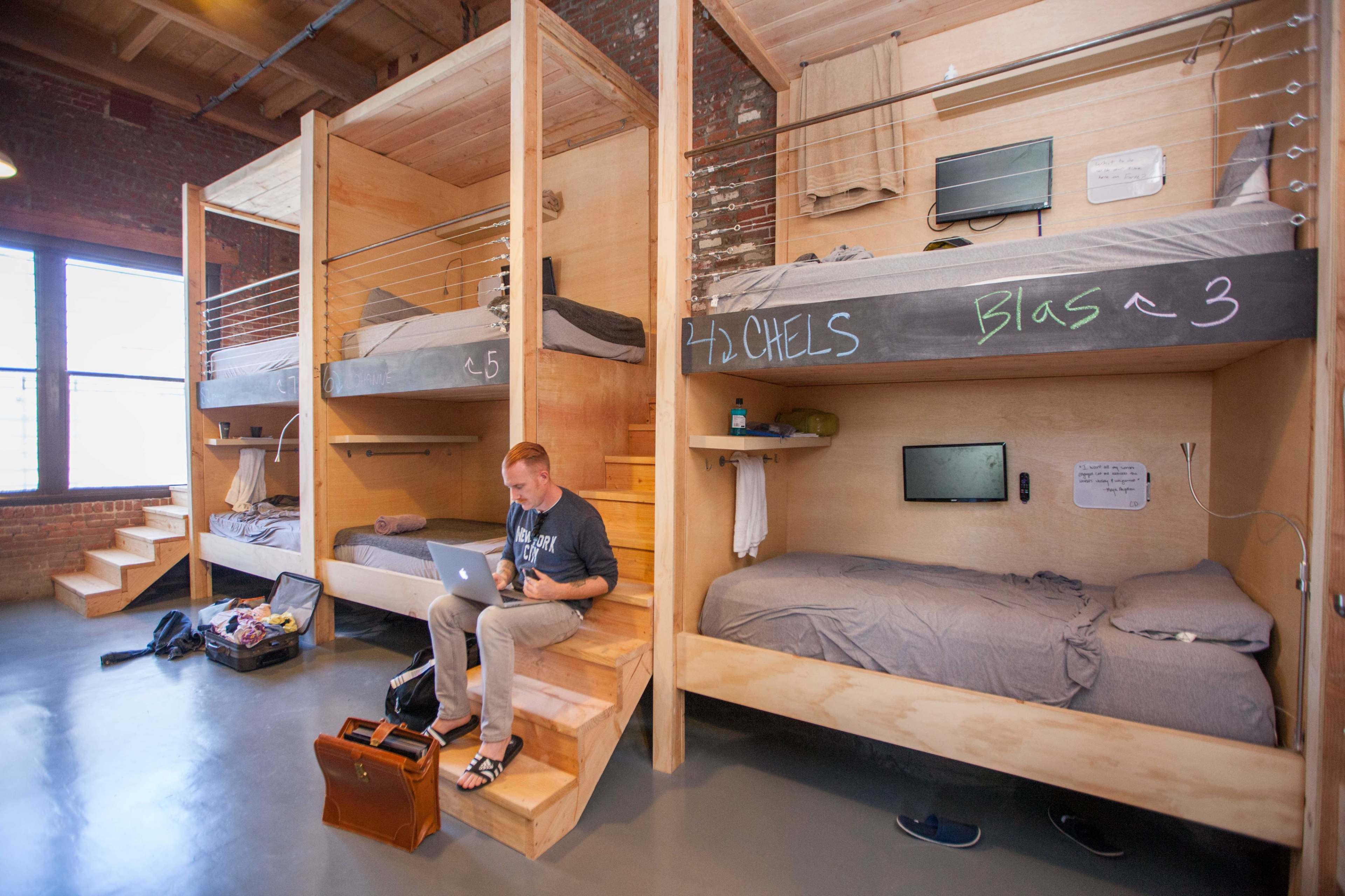 A modern dormitory-style room with wooden bunk beds and a man sitting on the stairs using a laptop.