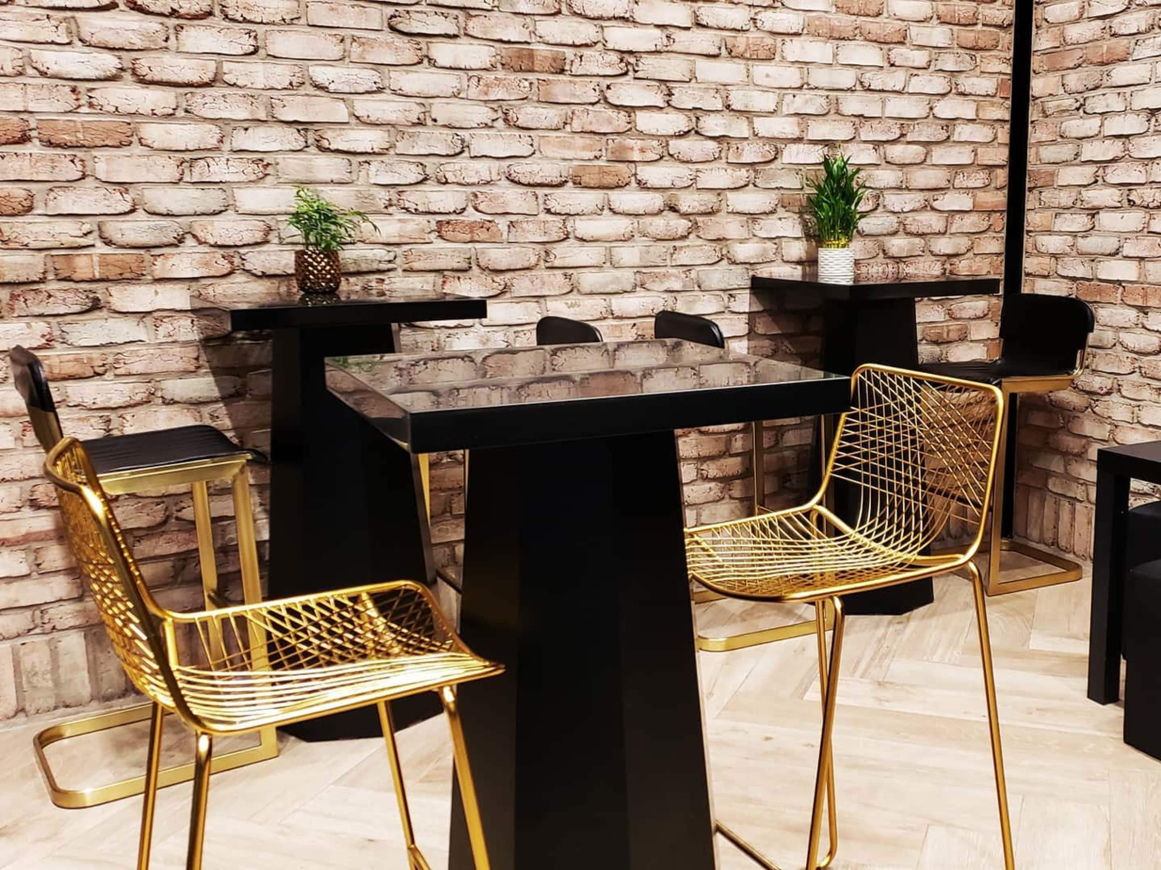 A modern interior space features black tables and gold wire chairs against a brick wall, with small potted plants on the tables.