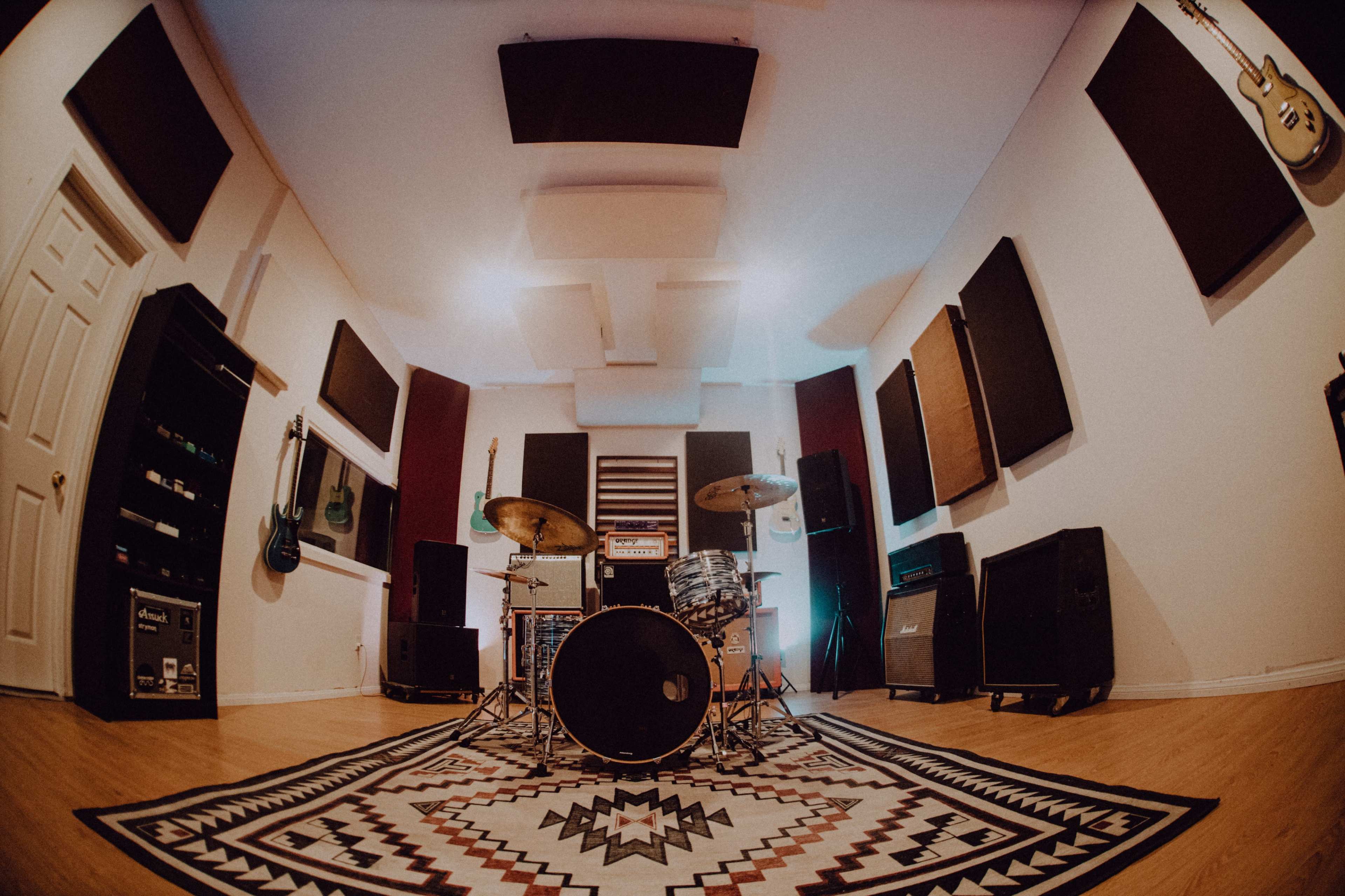 The image shows a music recording studio with a drum set, amplifiers, and soundproofing panels on the walls.