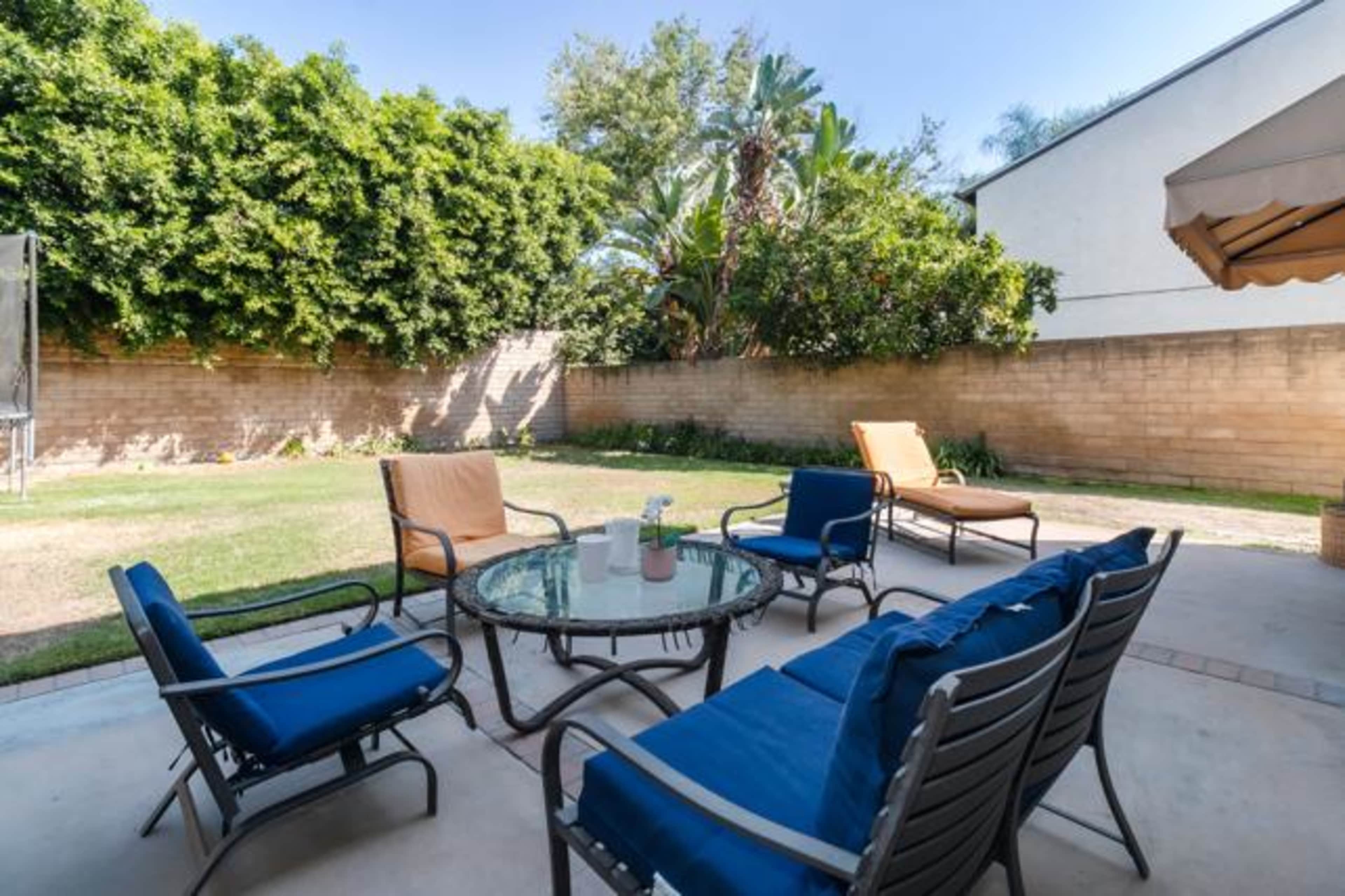 A backyard patio features a circular glass table surrounded by several lounge chairs, with a green lawn and shrubs in the background.