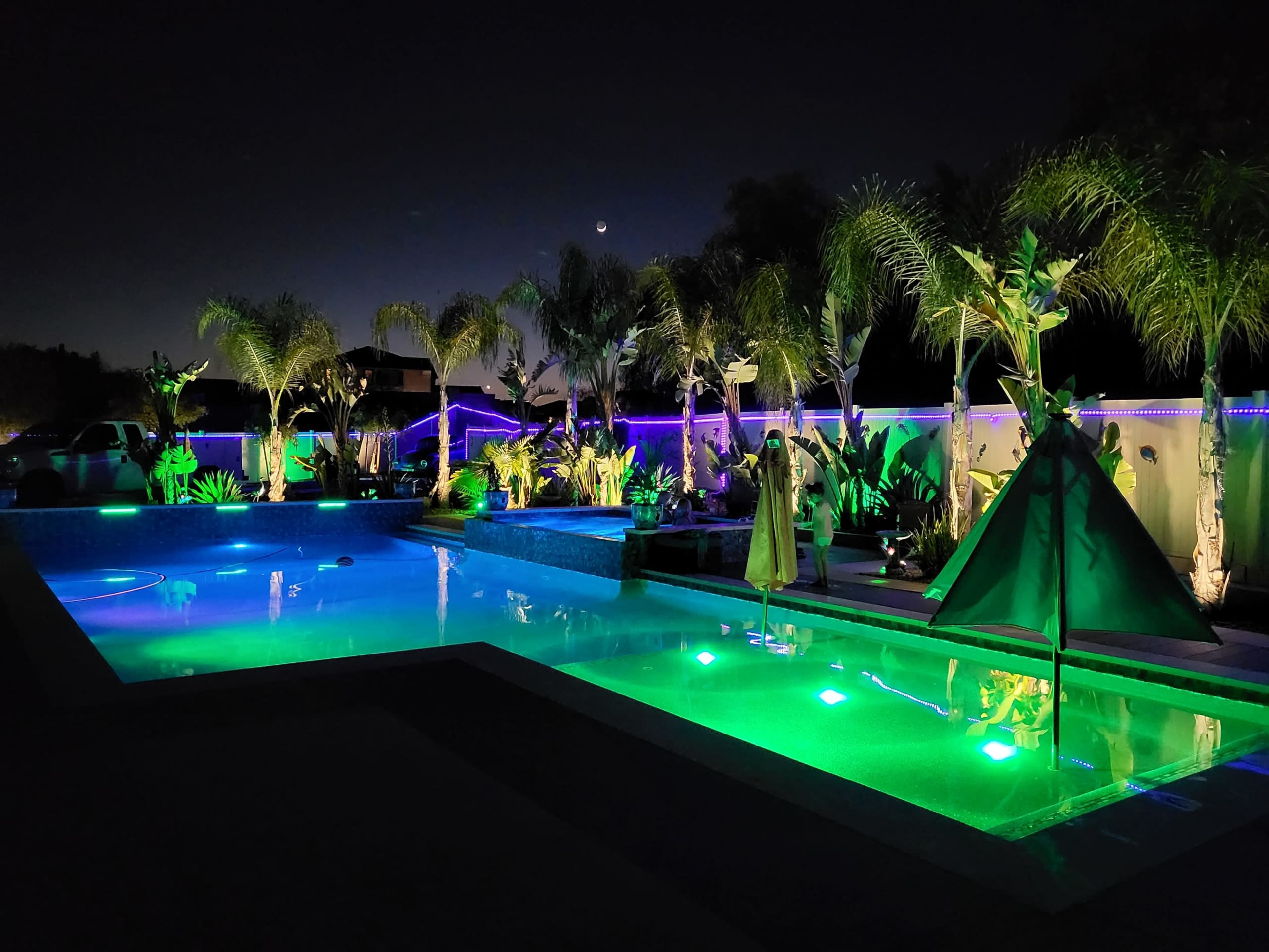 A well-lit backyard pool area featuring illuminated water, palm trees, and a tent-like structure, all under a night sky.