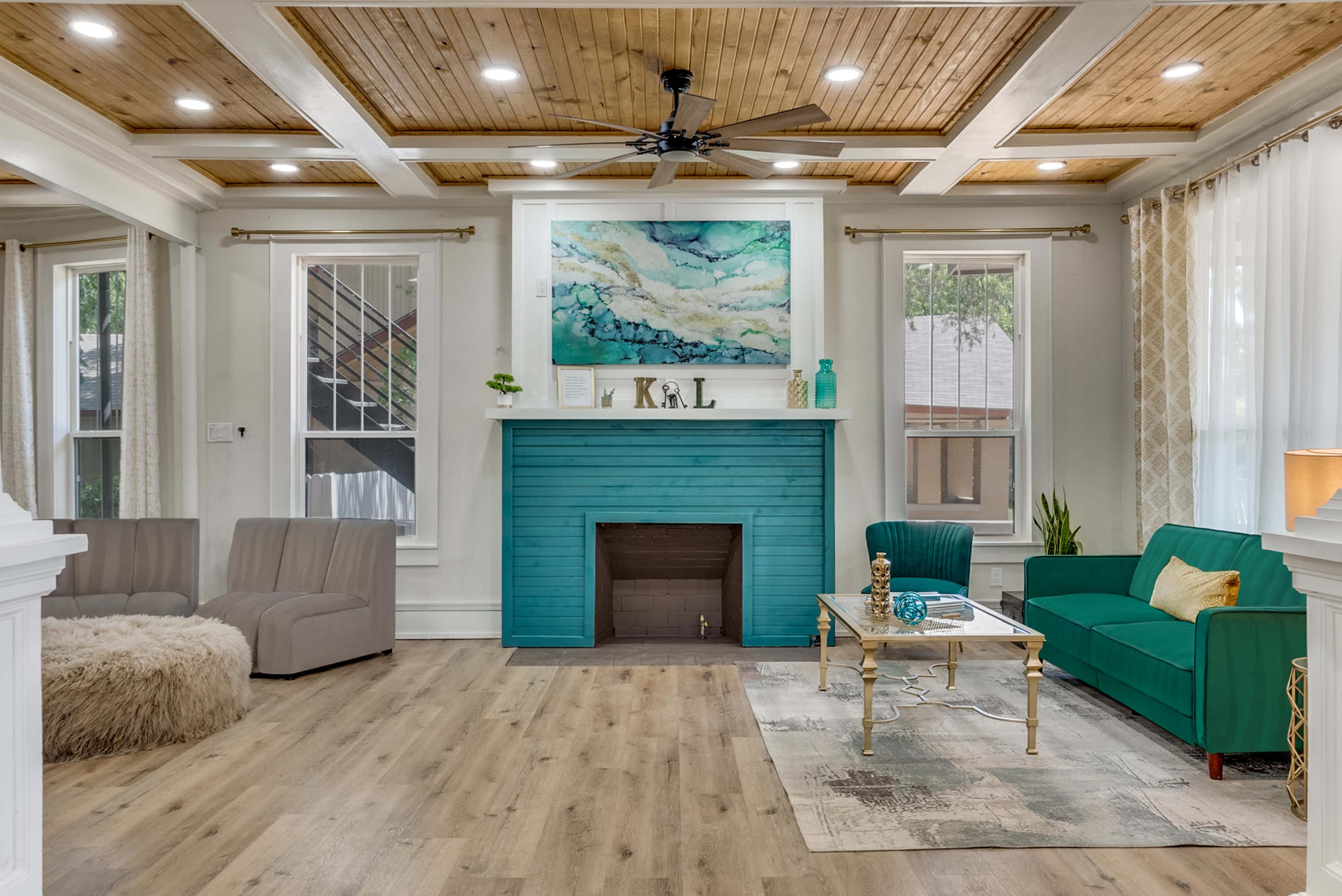 The image shows a modern living room featuring a teal fireplace, a green sofa, two gray armchairs, and large windows that allow natural light.