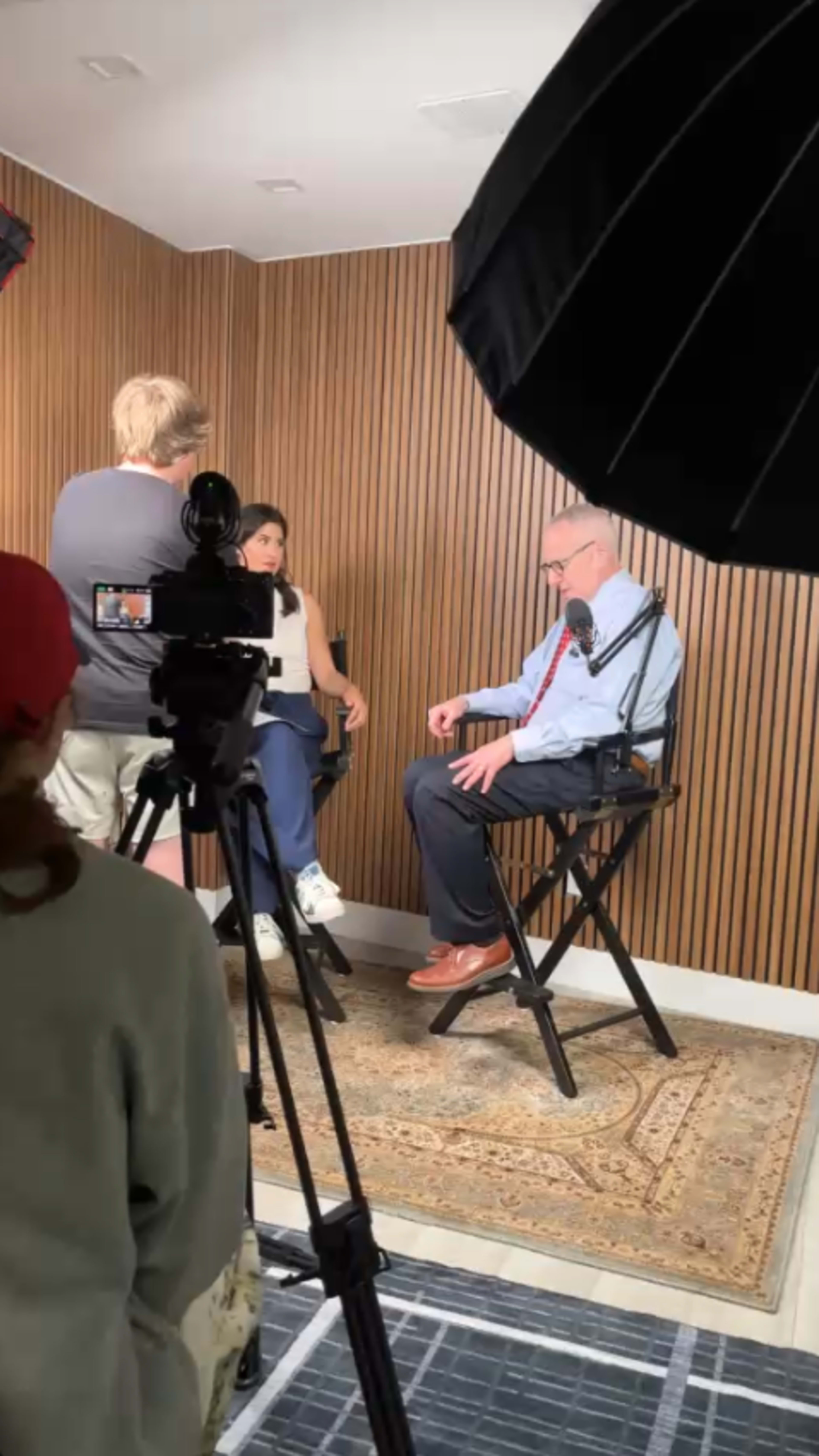 A person is being interviewed in a studio with wooden walls while a camera and crew are positioned around them.