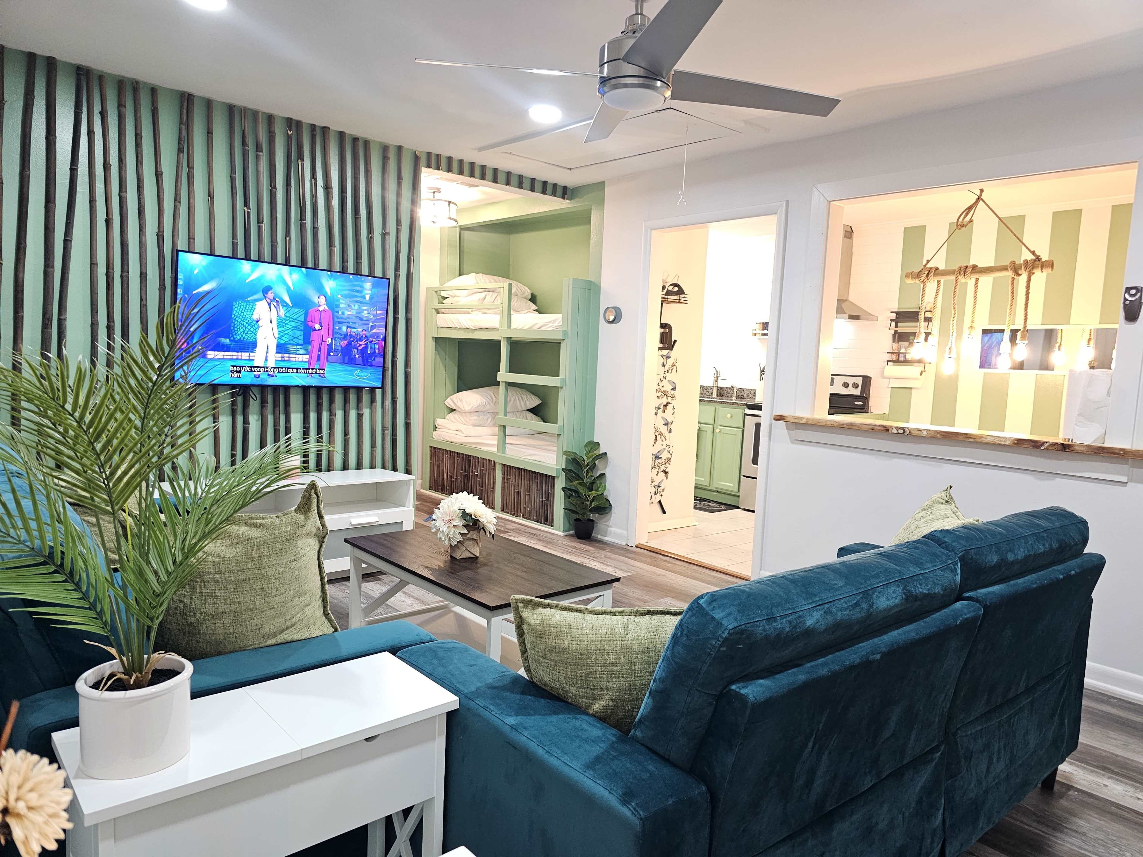 The image shows a modern living room with a teal sectional sofa, a television mounted on a patterned wall, and a view into a kitchen with green striped walls.