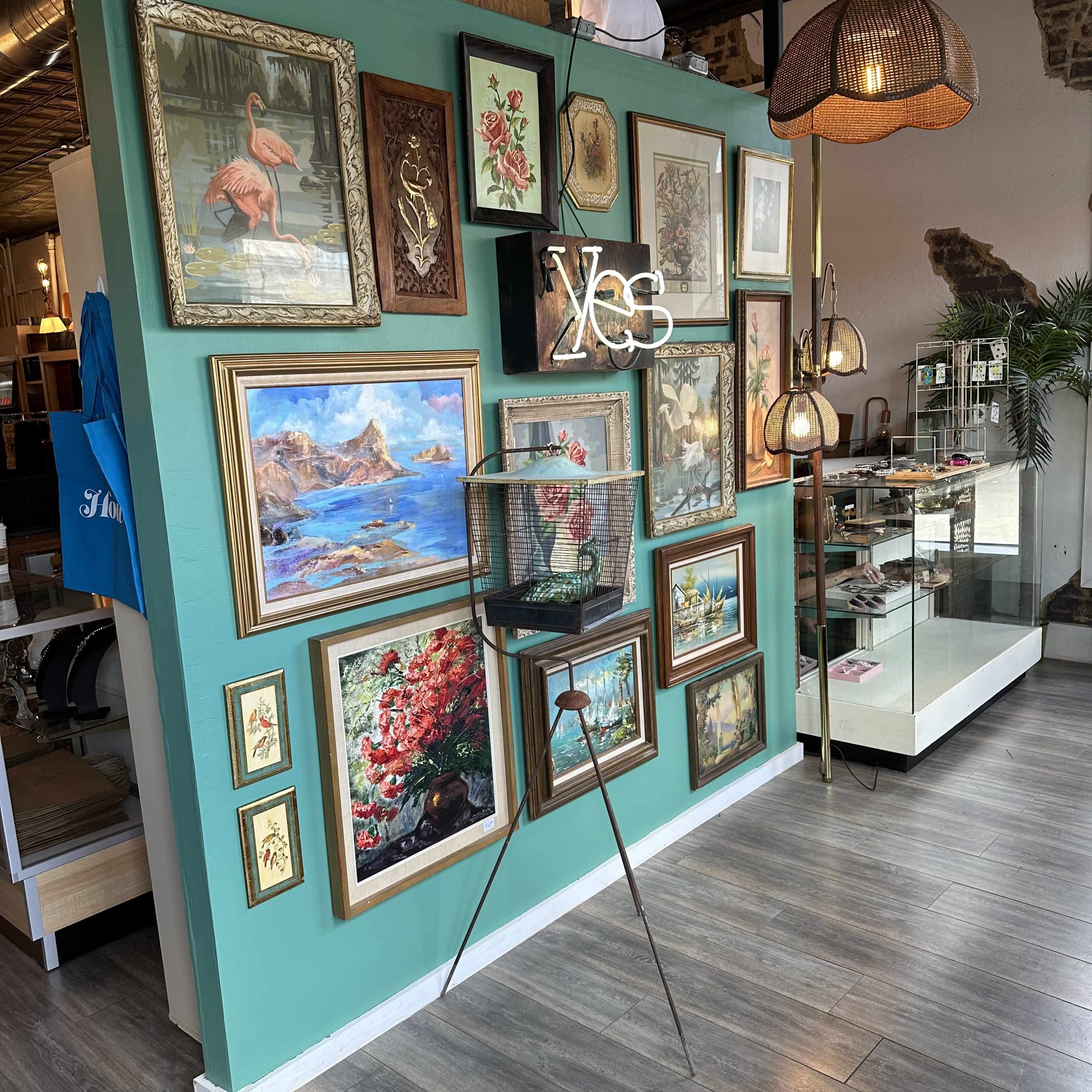 The image shows a decorated wall featuring various framed artworks, including landscapes and floral designs, along with a decorative stand and a light fixture in a retail space.